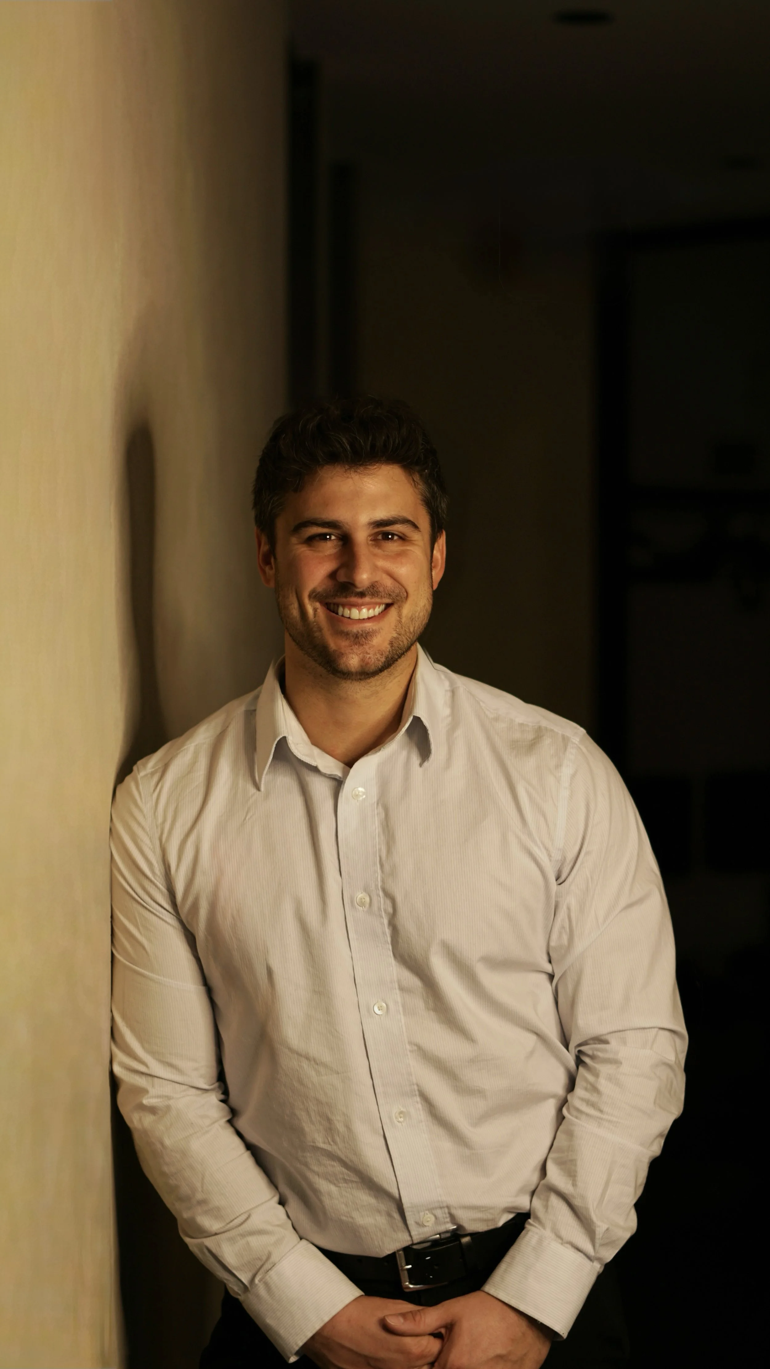 Courtier immobilier souriant portant une chemise blanche, debout à côté d'un mur beige dans une pièce sombre.