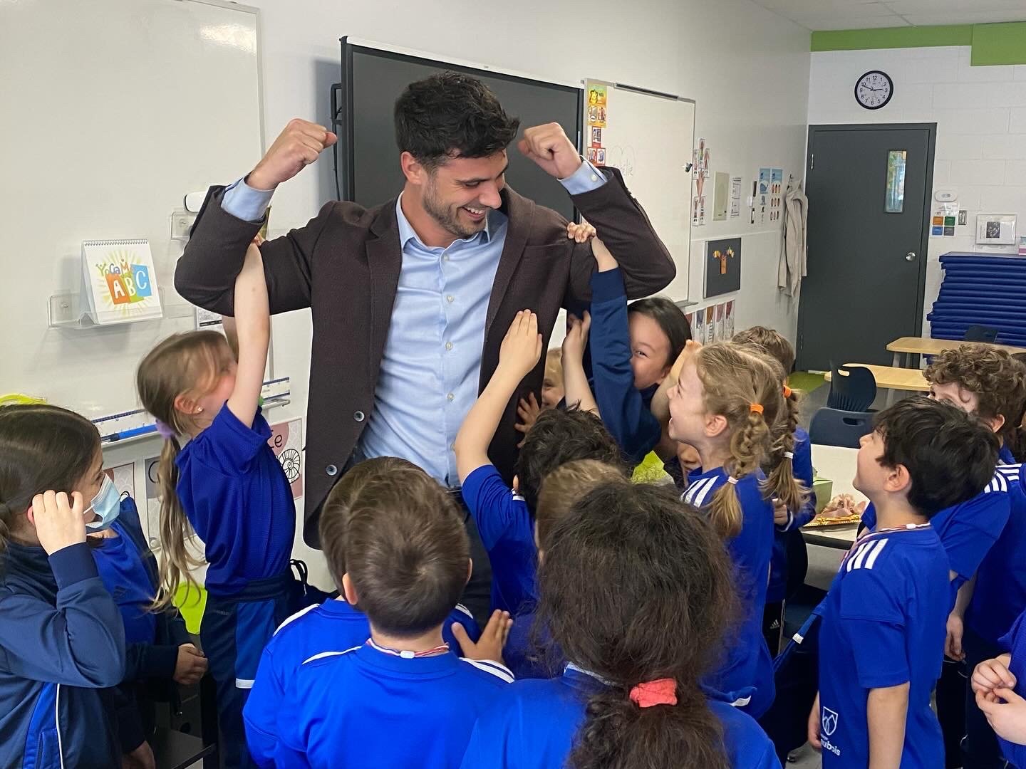 Un homme en costume saluant et levant ses bras en signe de victoire, entouré de plusieurs enfants de l'école portant des uniformes bleus, dans une salle de classe.