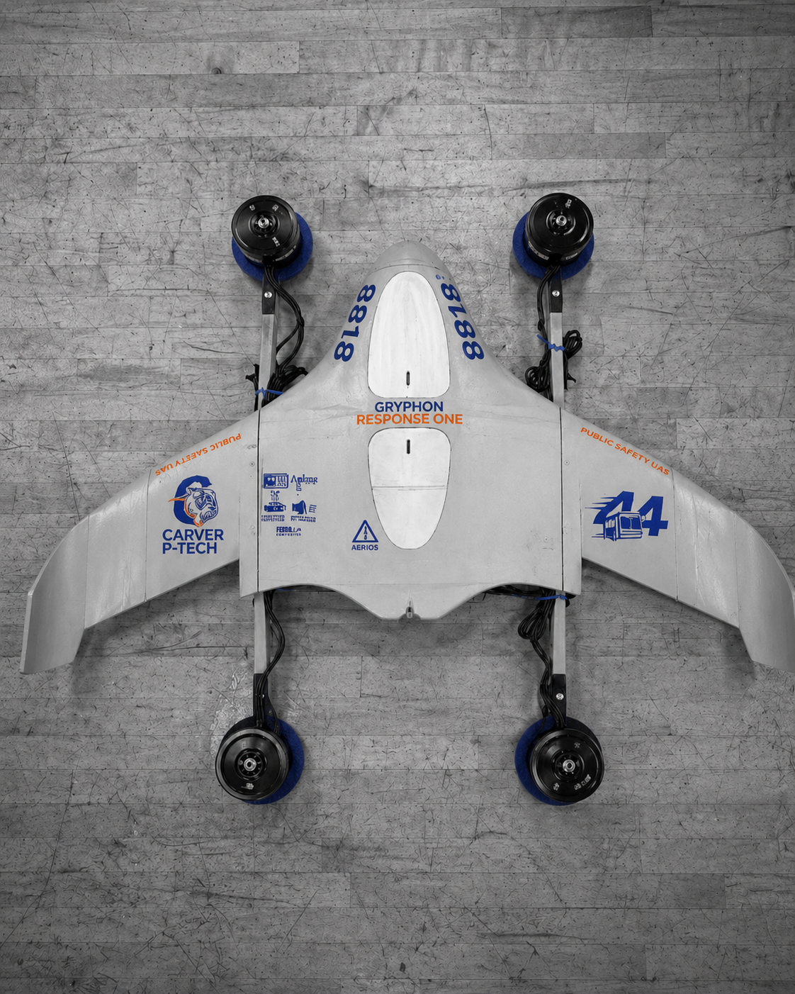 A white drone with four black wheels, blue protective covers on the wheels, and various blue and orange logos and text on its body, lying on a gray wooden floor.