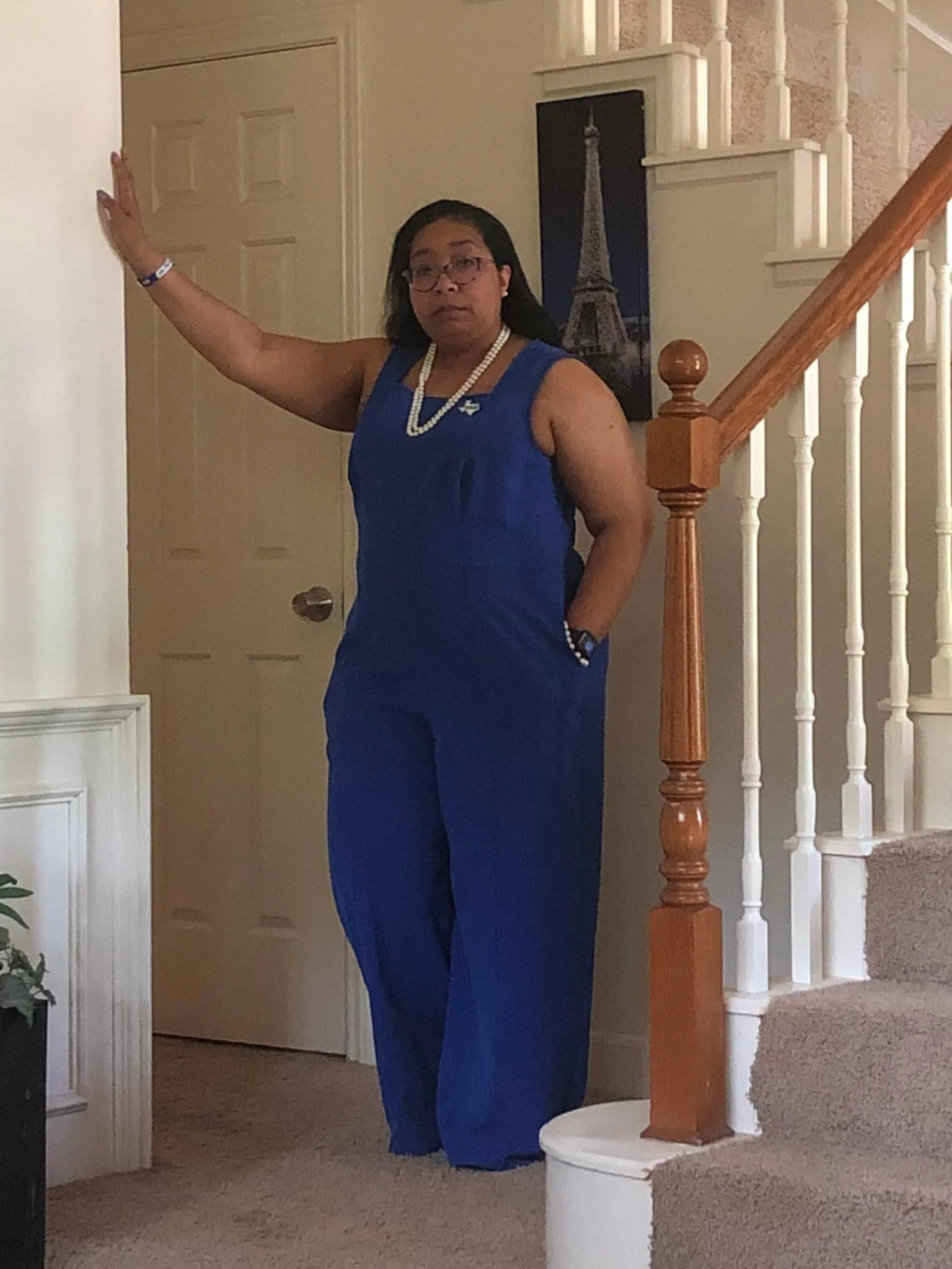 A woman wearing a blue dress, pearl necklace, and glasses standing inside a home near the staircase, with a picture of the Eiffel Tower on the wall behind her.