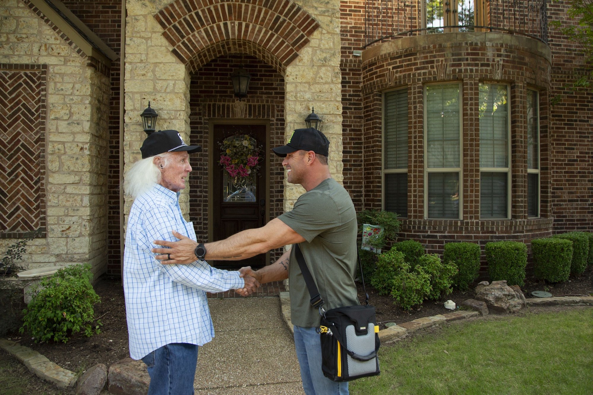 Two men shaking hands in front of a brick house with a front door adorned with a wreath. The older man has long white hair, and the younger man is wearing a cap and has a tool belt.