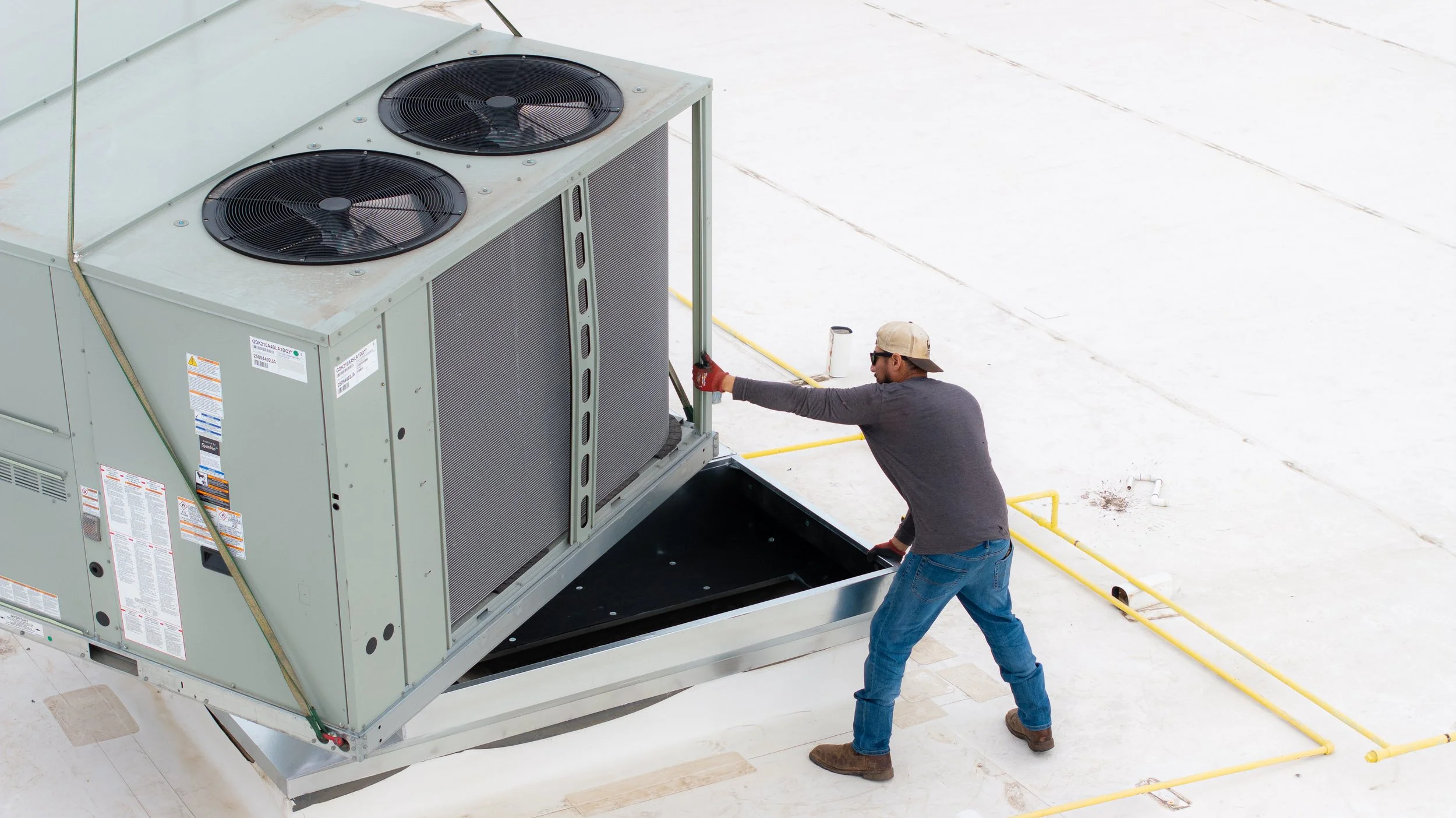 A man wearing a gray long-sleeve shirt, blue jeans, brown shoes, red gloves, a beige cap, and sunglasses is working on an HVAC unit on a flat, white rooftop of a building. He is holding a tool and appears to be repairing or maintaining the large gray equipment with two black fans on top.
