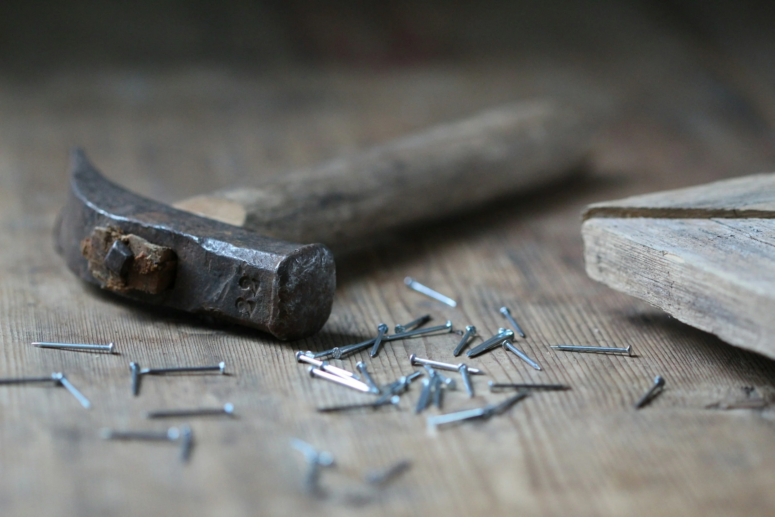 Close-up of a hammer head on a wooden workbench surrounded by scattered small metal nails and a piece of worn wood.