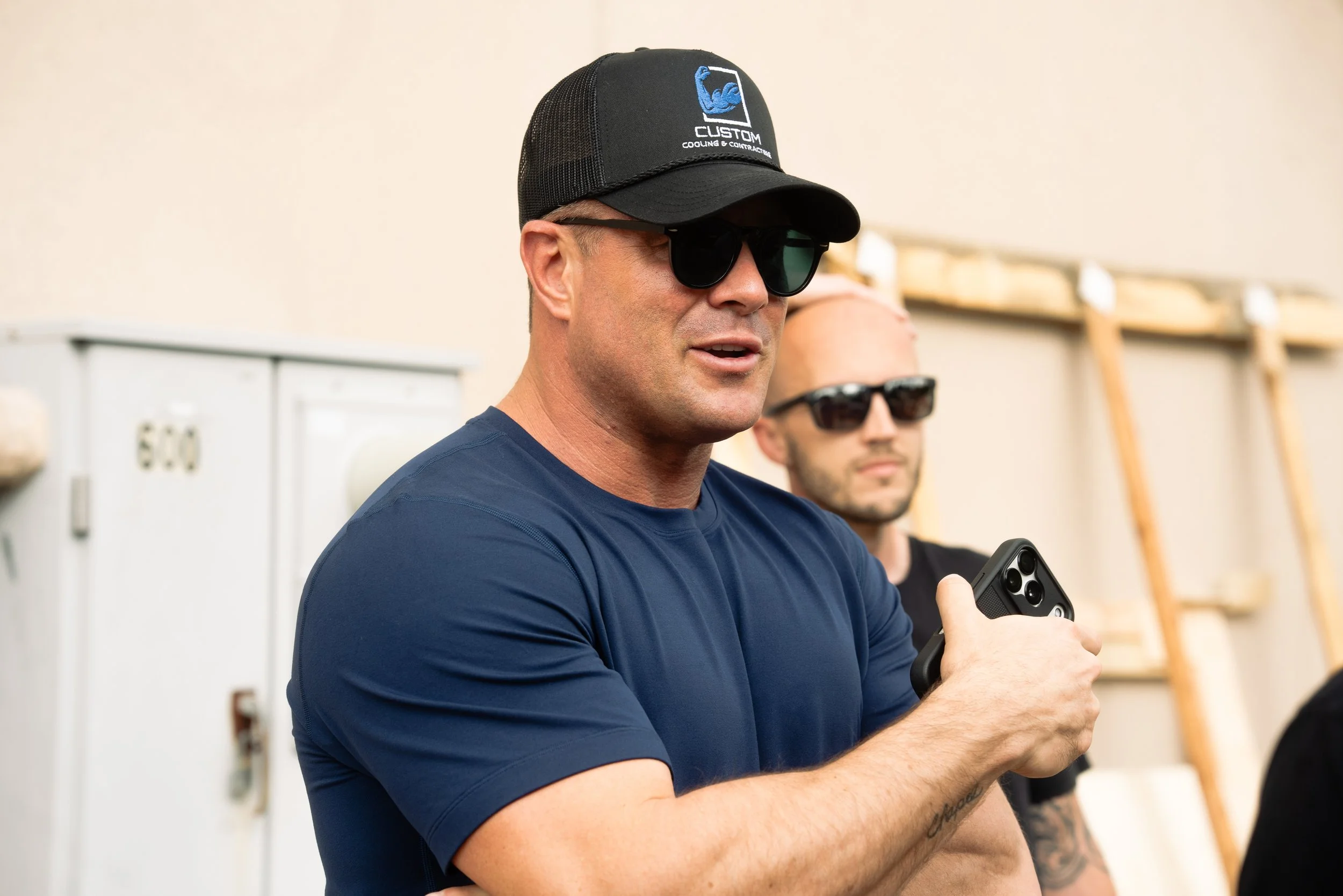 A man wearing sunglasses and a black trucker hat with a 'CUSTOM' logo, holding a smartphone, in a room with shelves and a metal cabinet in the background.