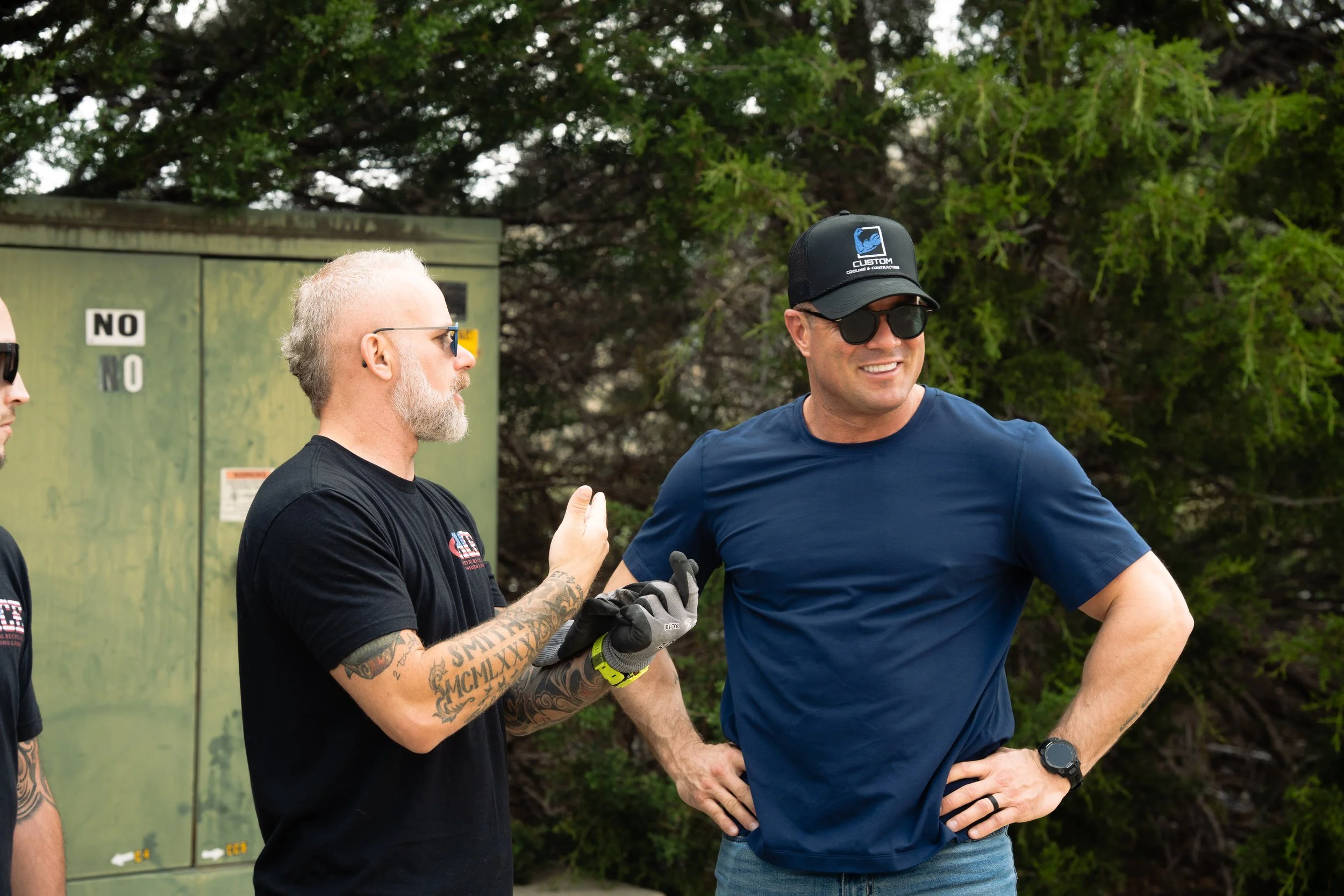 Two men talking outdoors, one wearing sunglasses, a black cap, and a blue shirt, the other with tattoos and glasses, in front of trees and a utility box.