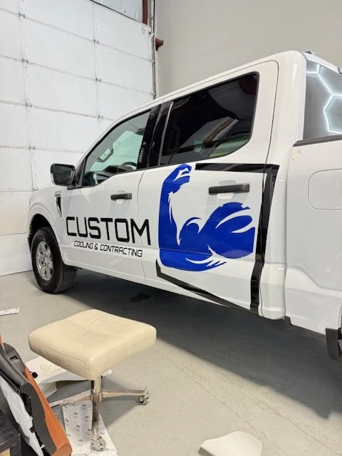 A white vehicle with a large blue graphic of a flexing arm and the text "CUSTOM COOLING & CONTRACTING" on the side, inside a garage or workshop.