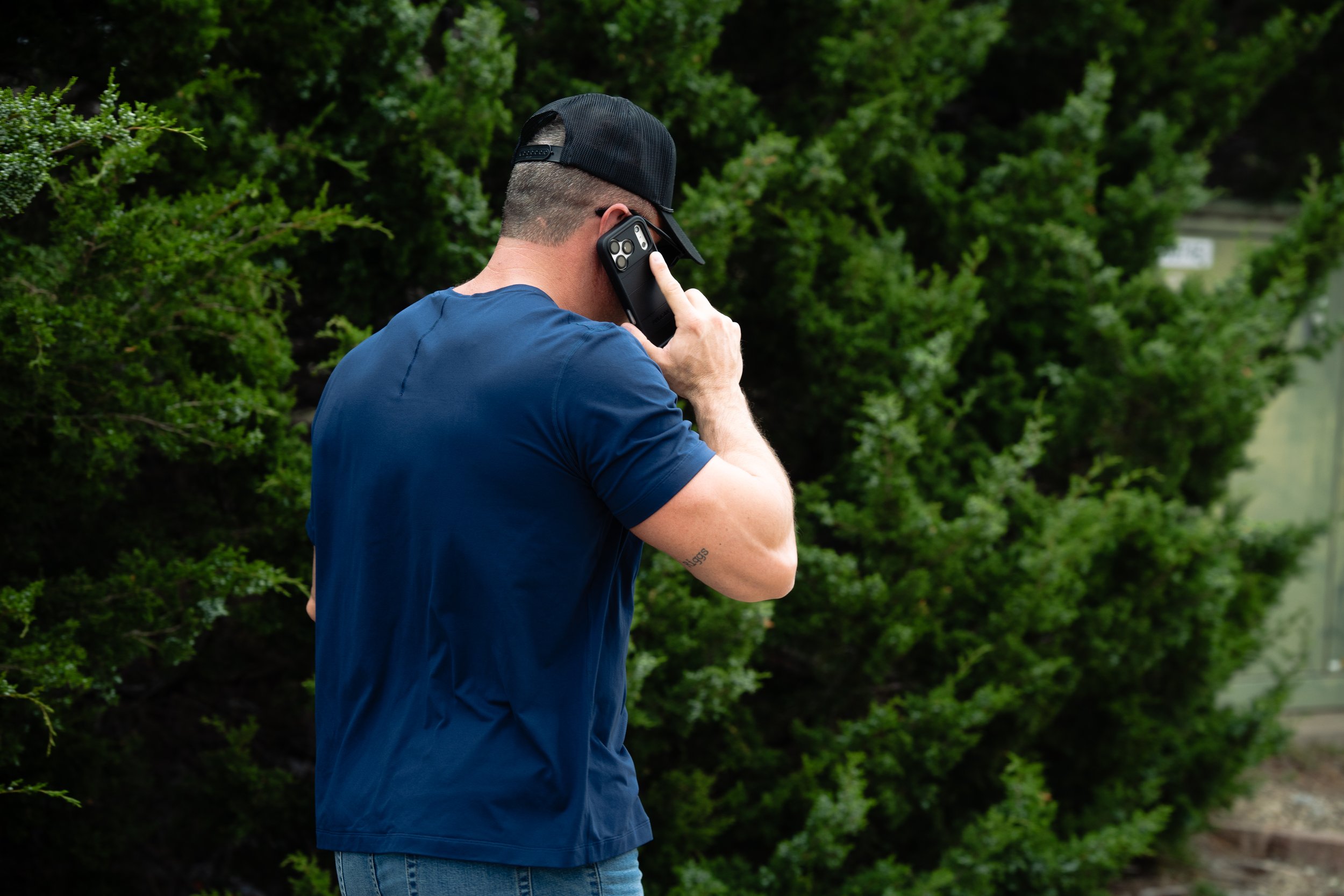 A man wearing a black cap, blue shirt, and jeans is talking on a smartphone outdoors near green bushes.