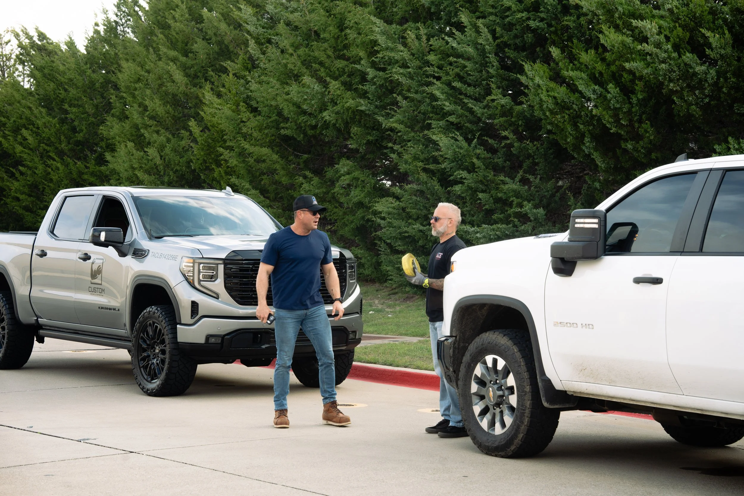 Two men standing near parked pickup trucks in a parking lot, with green trees in the background, having a conversation.
