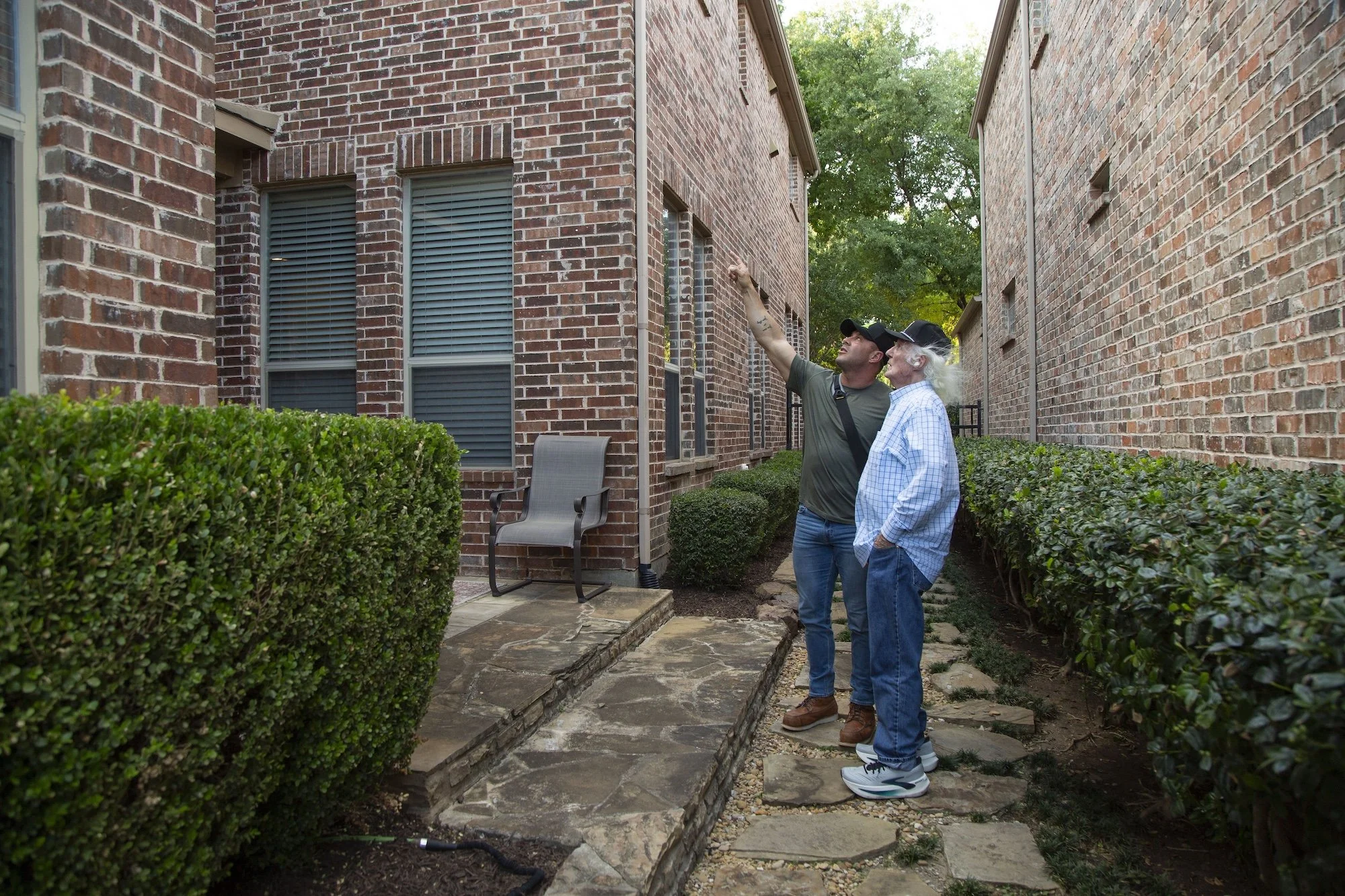 Two men standing outside between two brick buildings, one elderly with white hair and the other younger, pointing at the wall. They are on a stone paved pathway with greenery on sides and a chair in background.