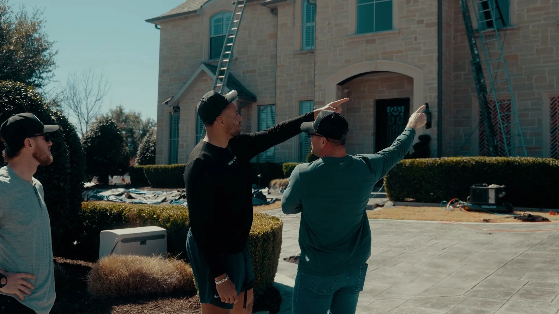 Three men discussing in front of a large house under construction, with ladders and construction equipment visible.
