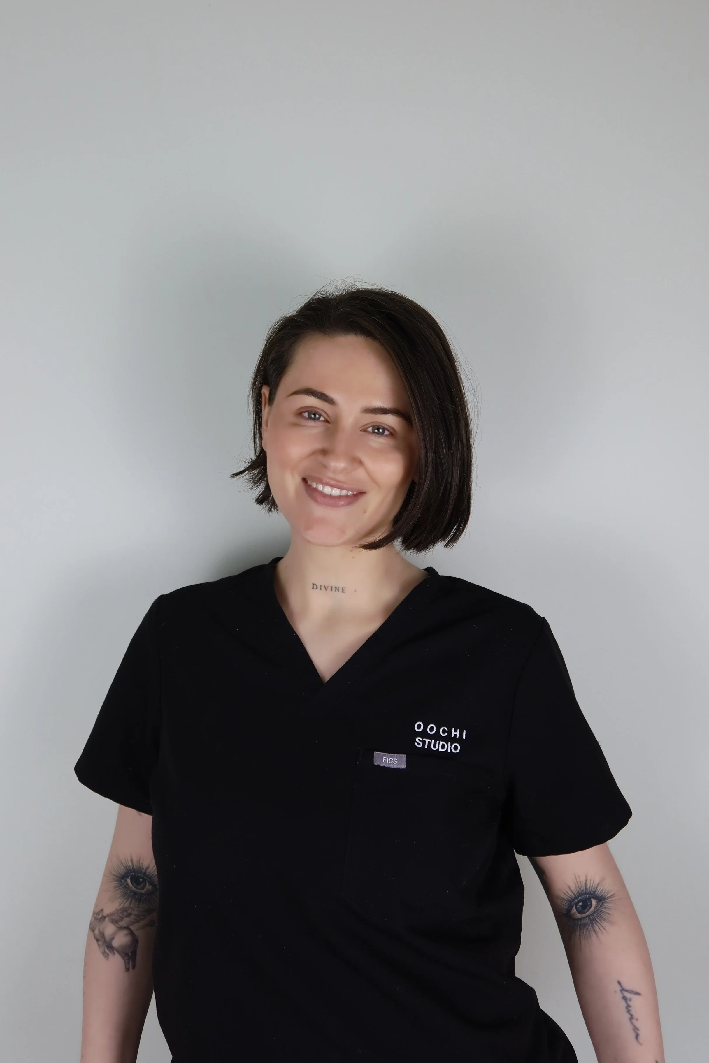 Bild av en kvinne med kort mørkt hår, smilende, kledd i svart uniform med tekst 'Oochi Studio' og 'Figs', har tatoveringer på underarmene med øye- og annet motiv.