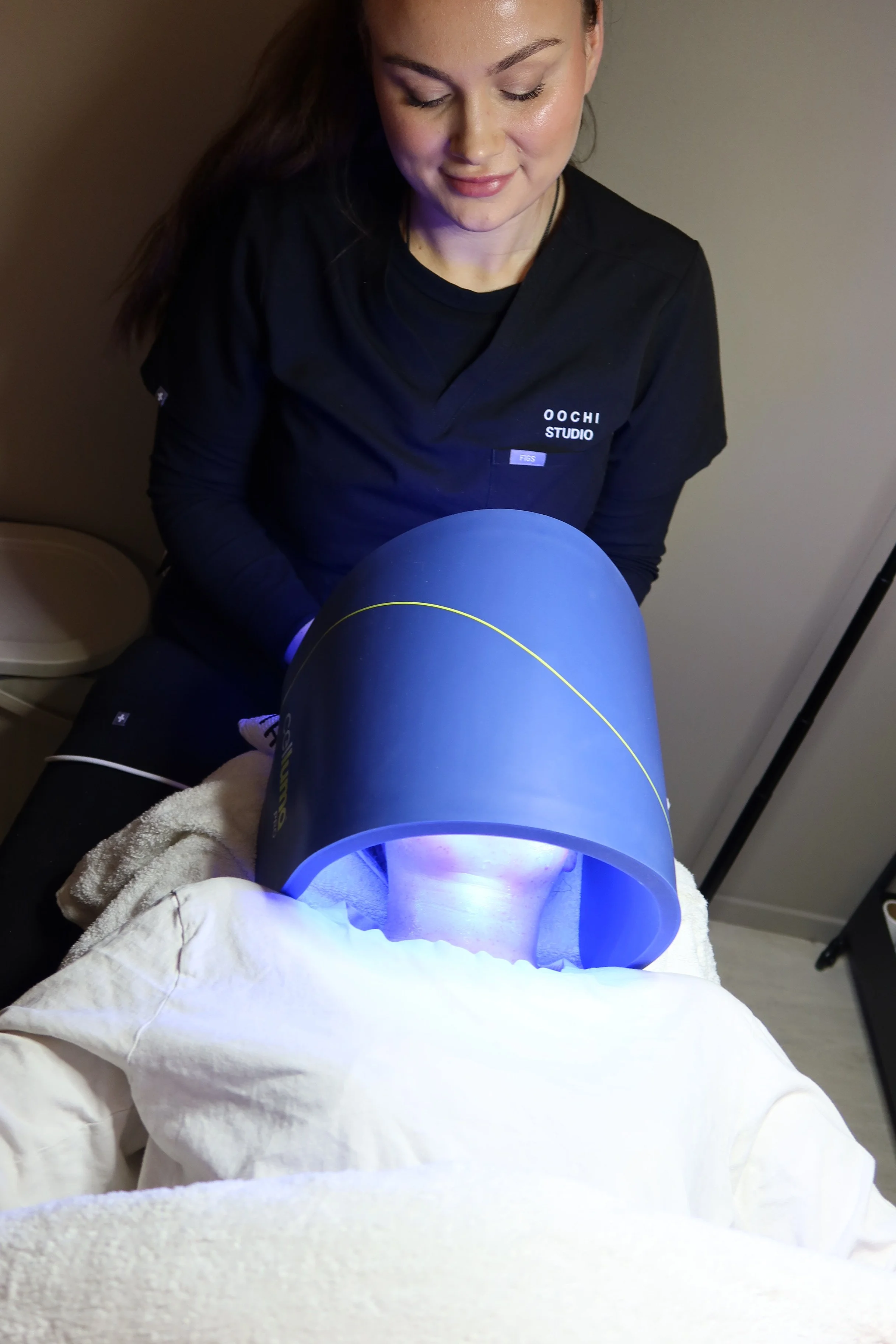 En kvinne med mørkt hår som utfører en ansiktsbehandling under en blå UV-lyslampe, iført svart uniform med teksten "Oochi Studio"