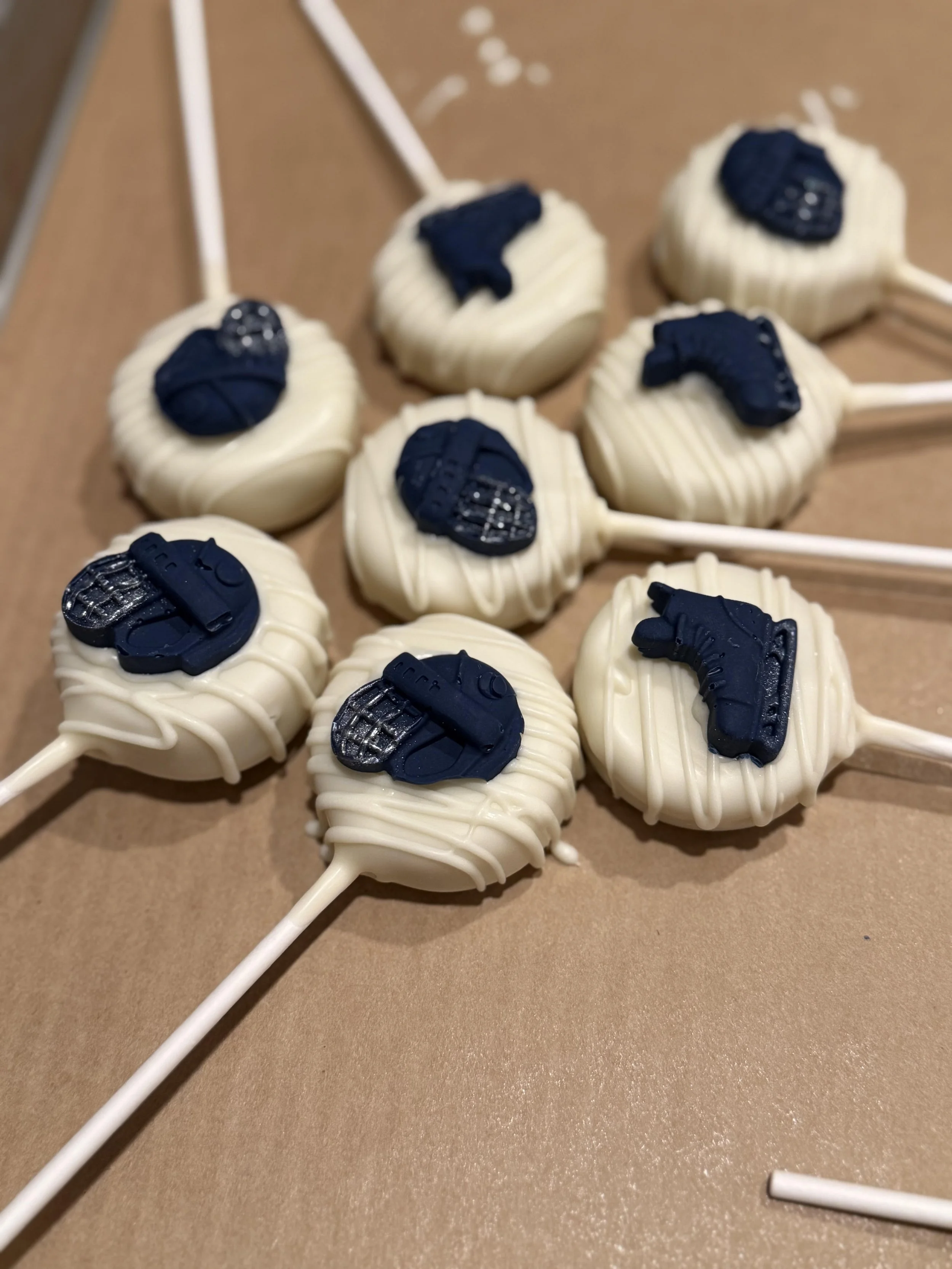 Cake pops with white chocolate coating and blue decorative hockey shapes on sticks, arranged on a brown surface.