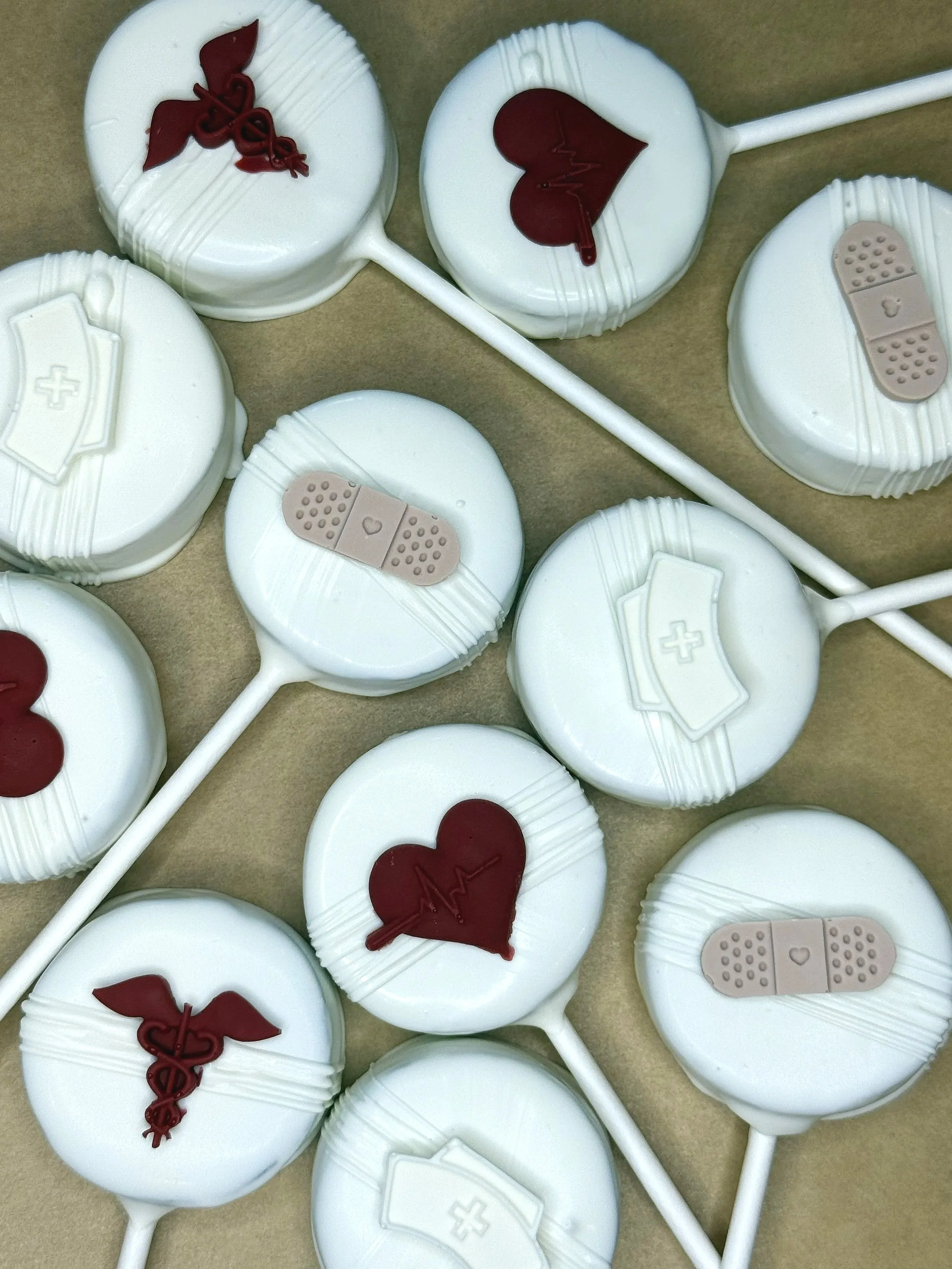 A collection of white lollipops with medical-themed decorations on top, including a red heart with an EKG line, a red caduceus symbol, a beige bandage, and a white medical cross.