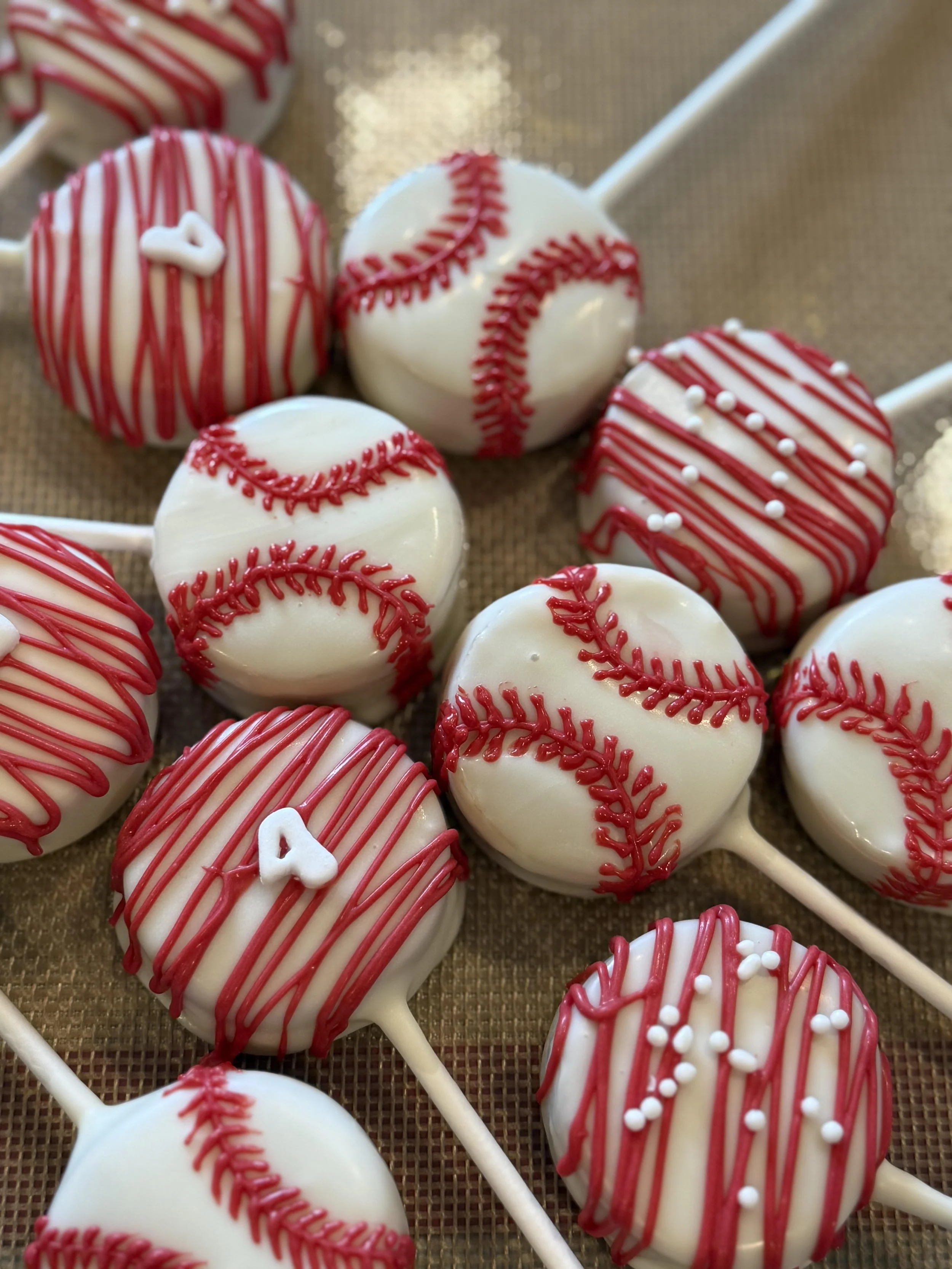 Assorted baseball-themed cake pops decorated with red and white icing, including red stitching and small white sprinkles, on sticks placed on a textured surface.