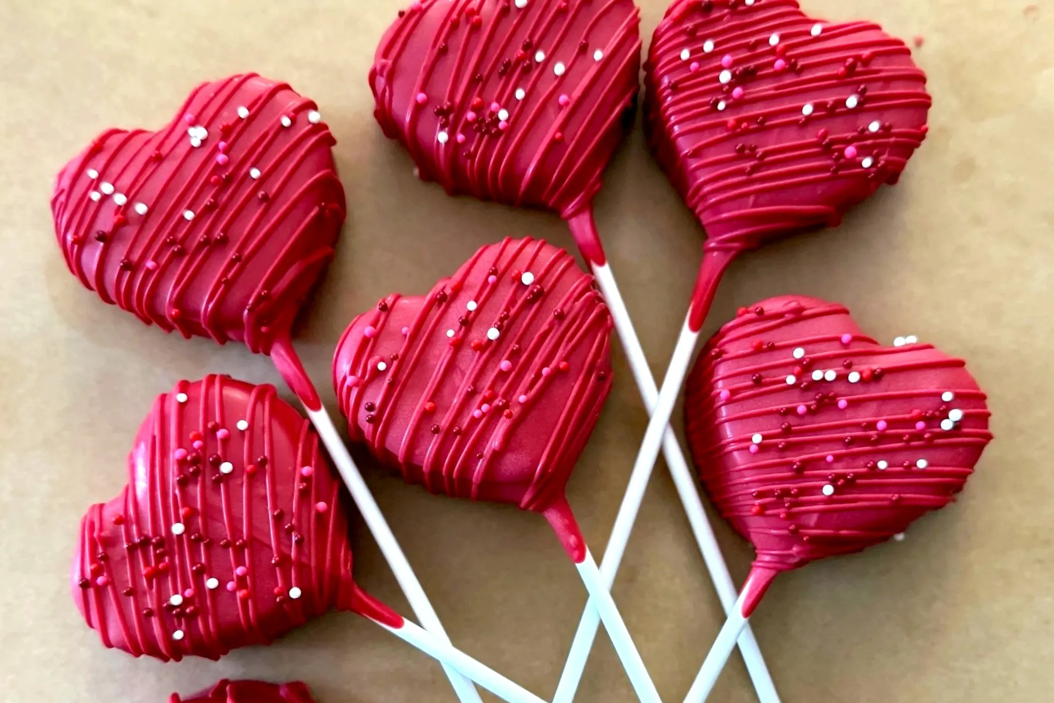 Six heart-shaped red lollipops decorated with pink, white, and red sprinkles on a beige background.