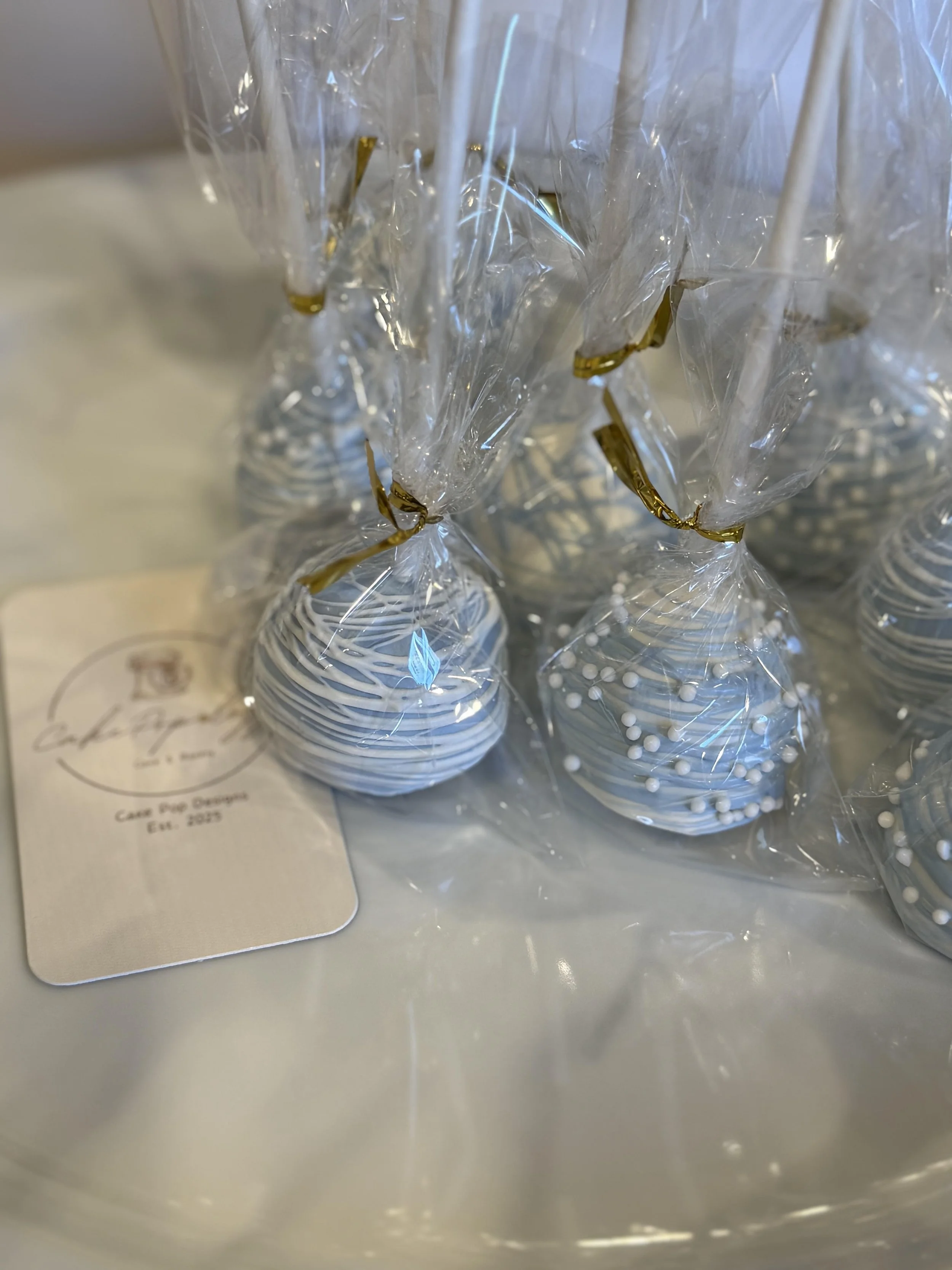 Close-up view of white and blue cake pops with a shiny gold wrapper, enclosed in clear plastic bags, resting on a white surface with a partially visible note beneath.