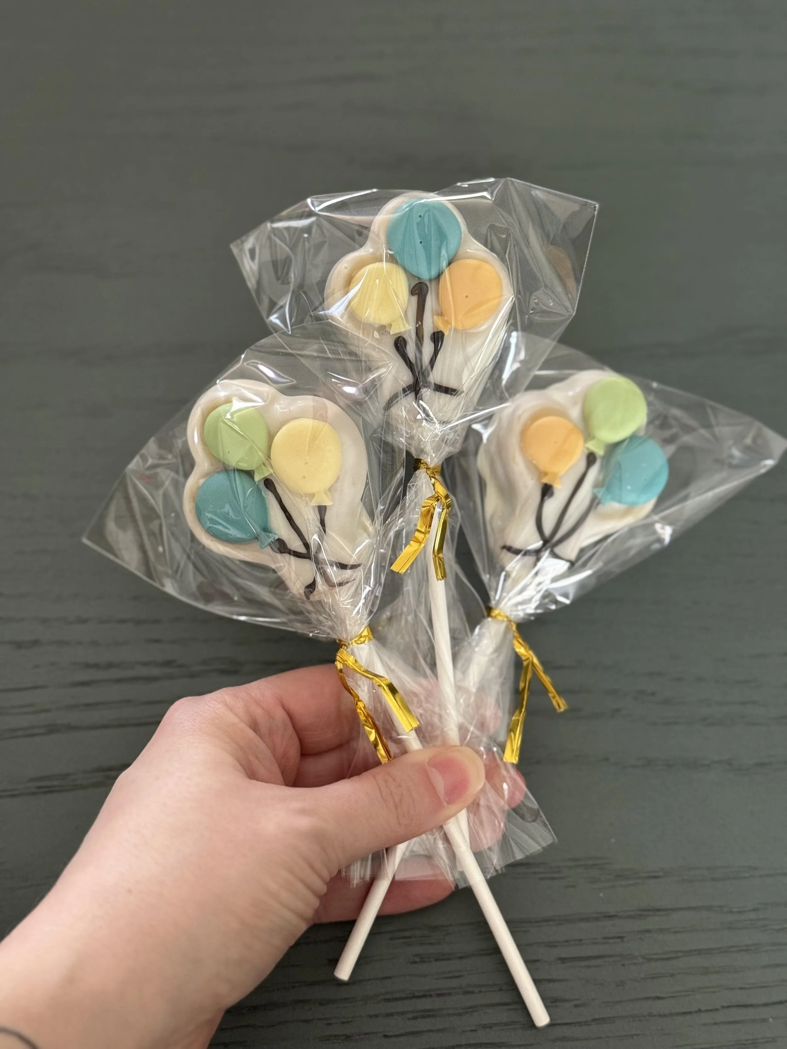 Hand holding three decorated cake pops shaped like balloons, wrapped in clear plastic with gold ties, on black sticks.