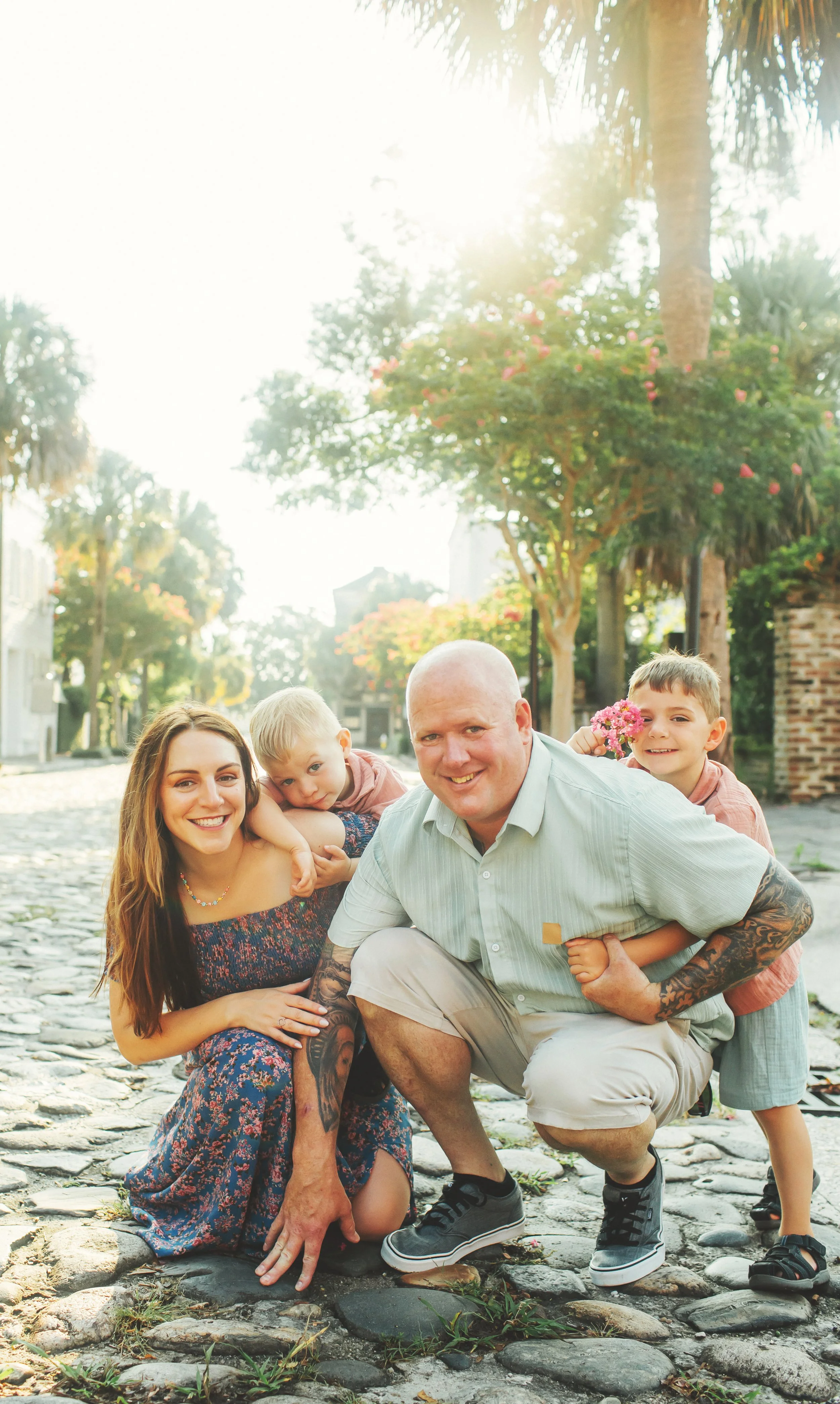 Family of four smiling and posing outdoors on a cobblestone street with trees and historic buildings in the background, sunny weather.