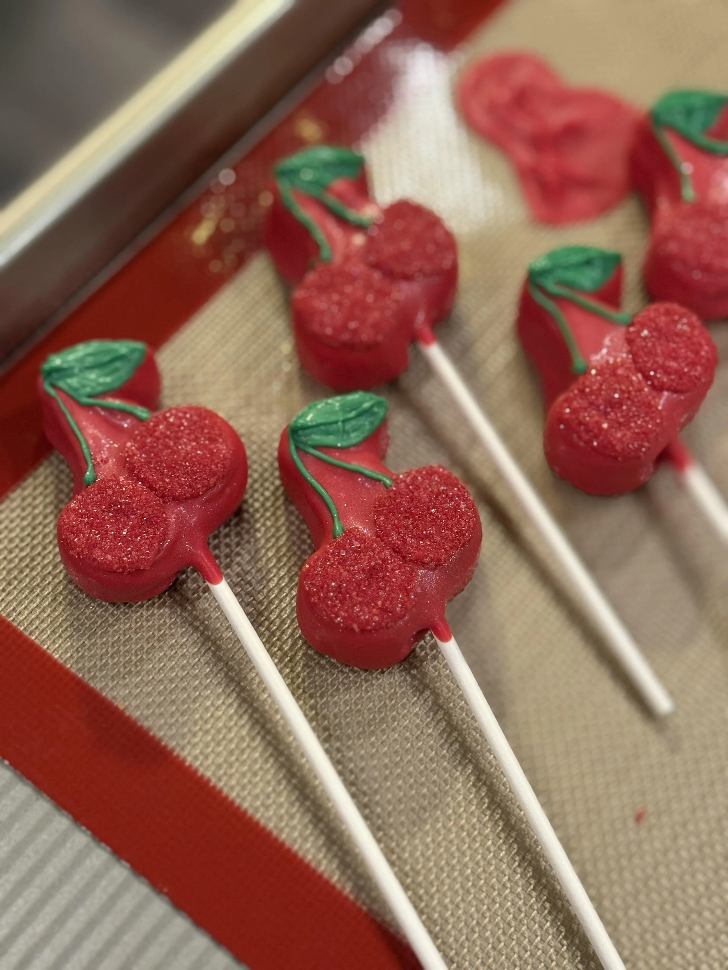 Close-up of red cherry-shaped cake pops decorated with green leaves, placed on a baking mat with a red edged baking sheet.