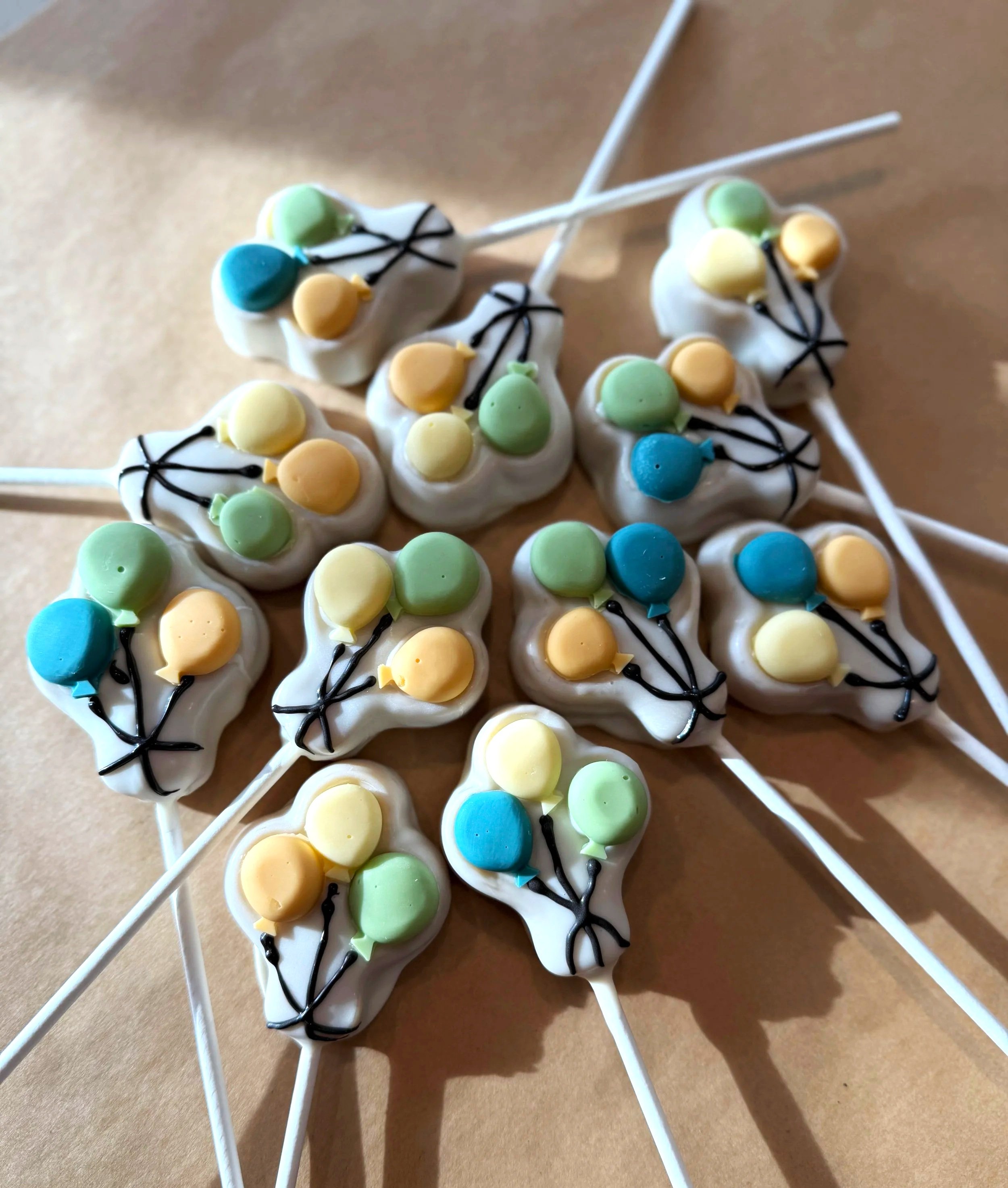 Colorful cake pops decorated with white icing, green, yellow, and blue candy balloons, and black icing lines, arranged on white sticks on a cardboard background.