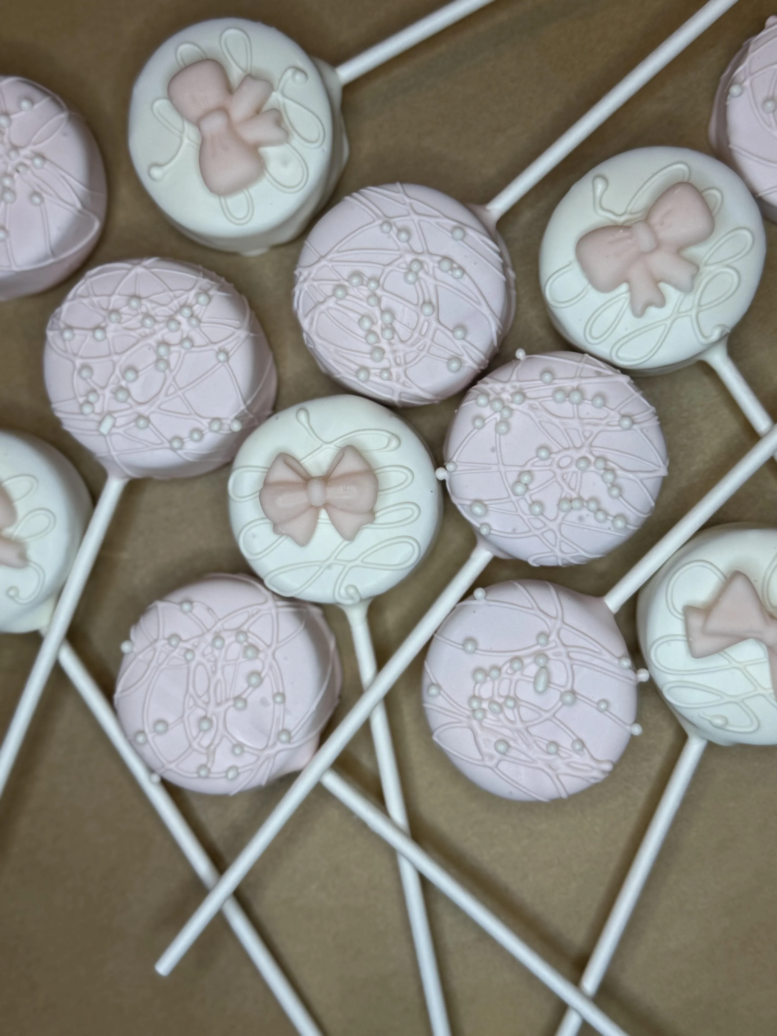 Close-up of pastel-colored decorated cake pops with white and pink icing, some topped with delicate white pearls and fine white lines, arranged on a brown surface with white sticks.