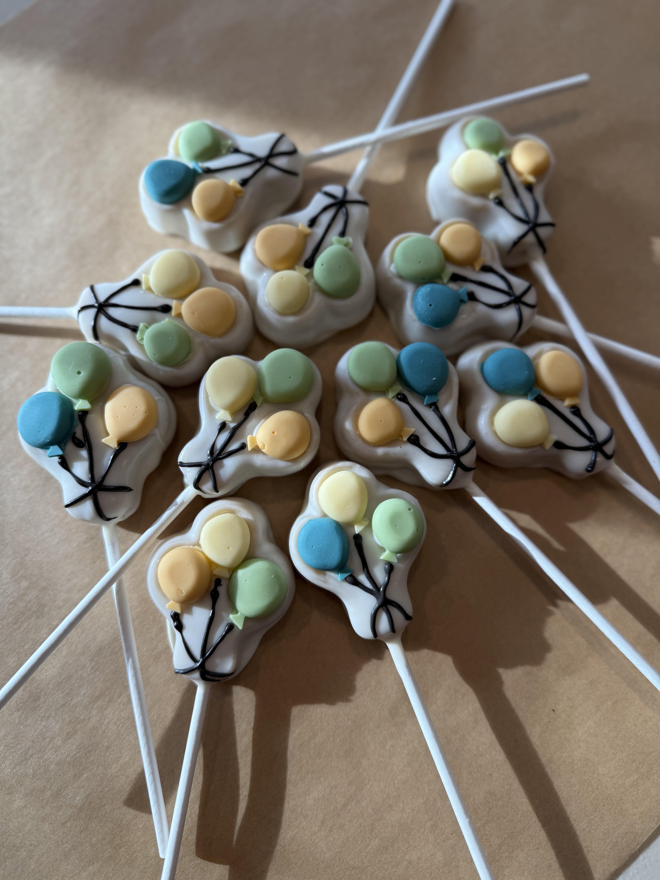 Assorted colorful cake pops with pastel yellow, green, and blue toppings, decorated with black lines, placed on white sticks and arranged on a brown surface.