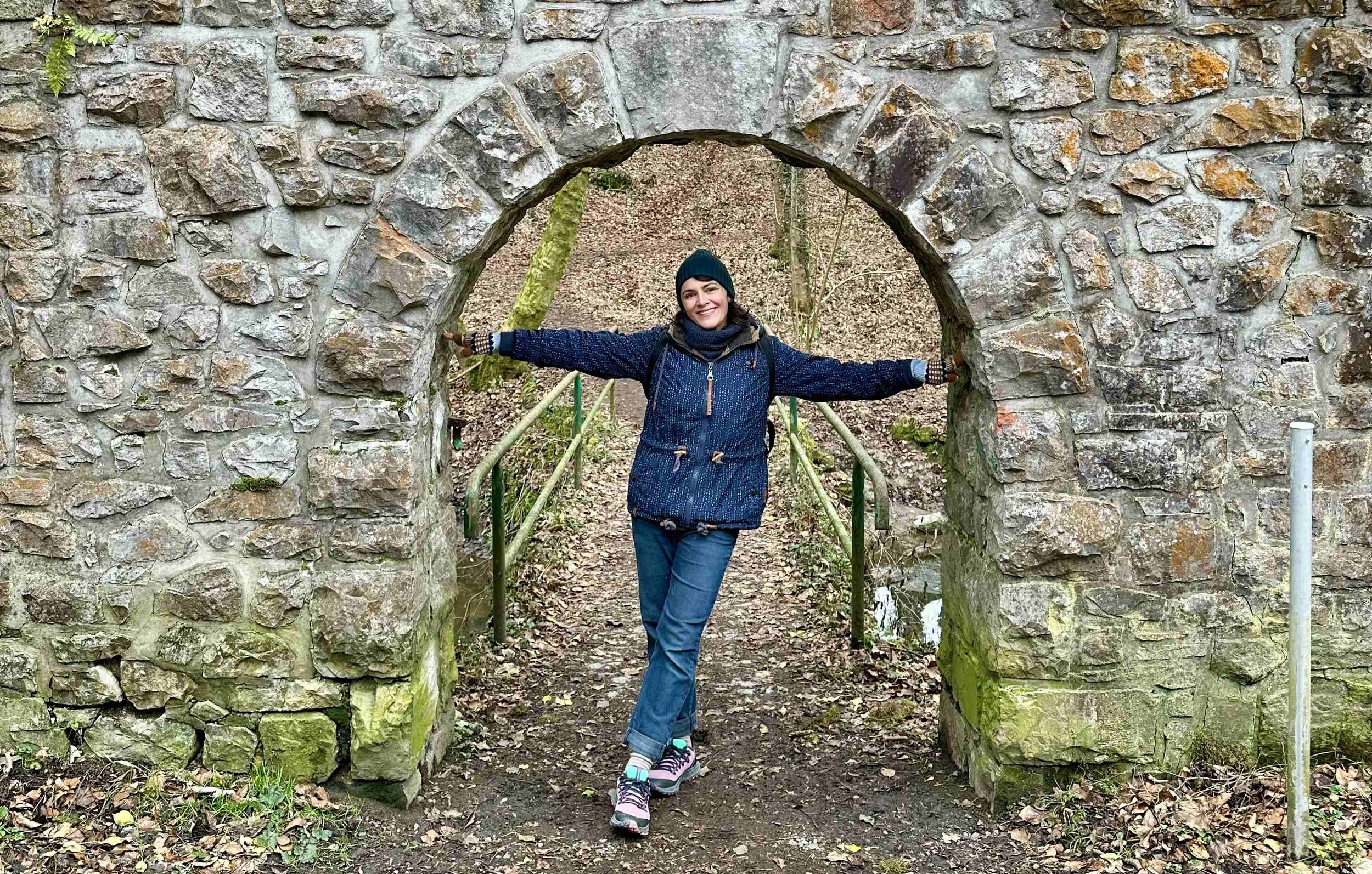 Devon Holzwarth standing beneath a stone archway in a wooded setting during an outdoor walk.