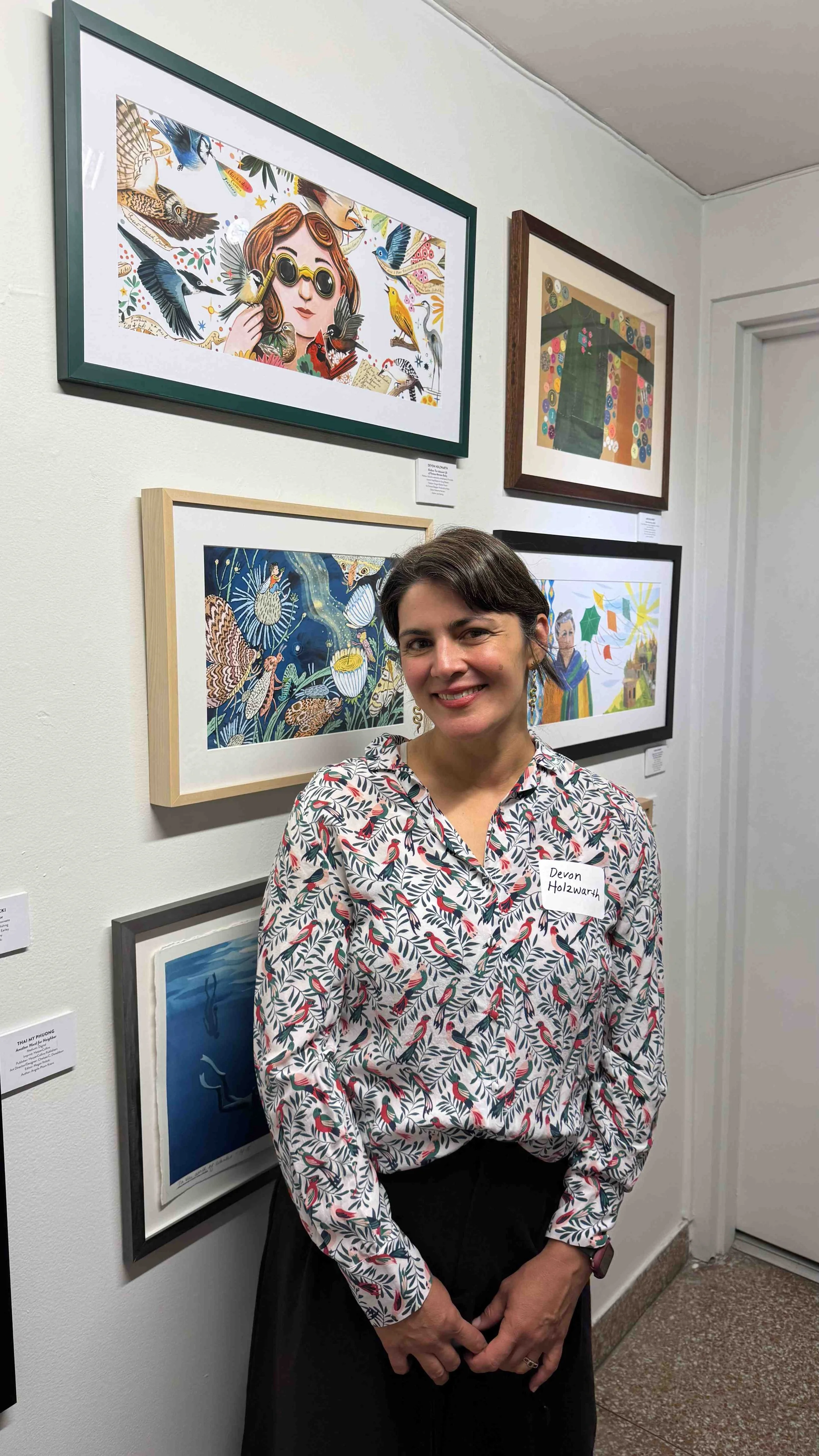 Devon Holzwarth standing beside framed artwork displayed in a gallery exhibition of her illustrations.