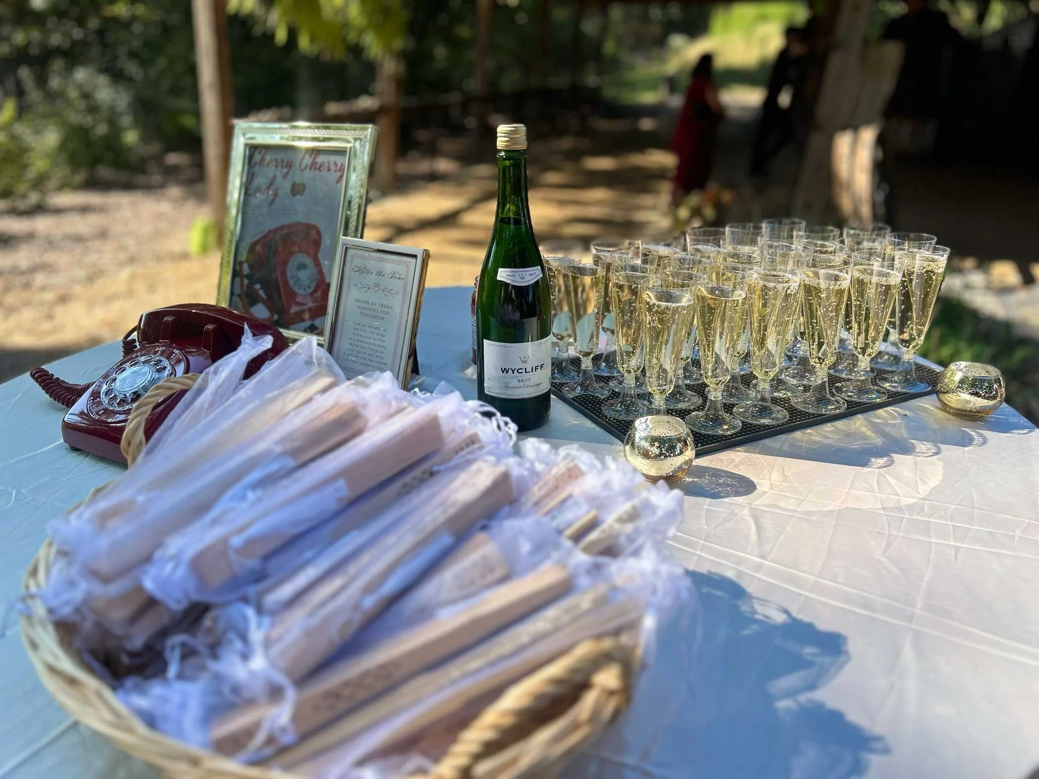 Table with a basket of bread, a wine bottle, several glasses of champagne, and framed signs at an outdoor event with trees and people in the background.