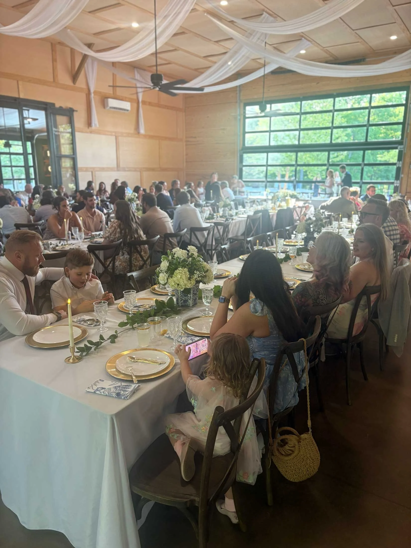 People gathered for a wedding reception or celebration in a wooden hall with large windows, decorated with draped fabric on the ceiling and floral centerpieces on the tables.