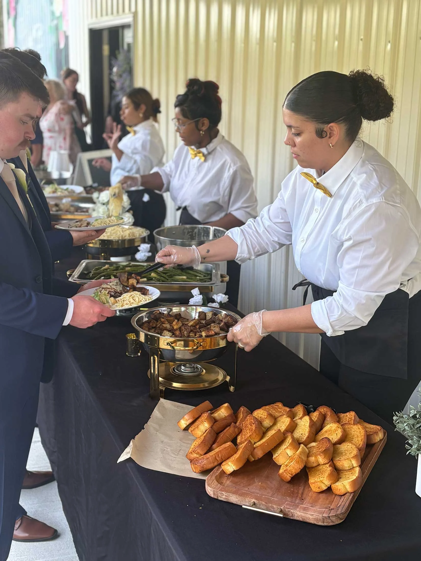People in line at a buffet-style food table with bread, pasta, and cooked vegetables, served by staff members in white shirts with yellow bow ties.