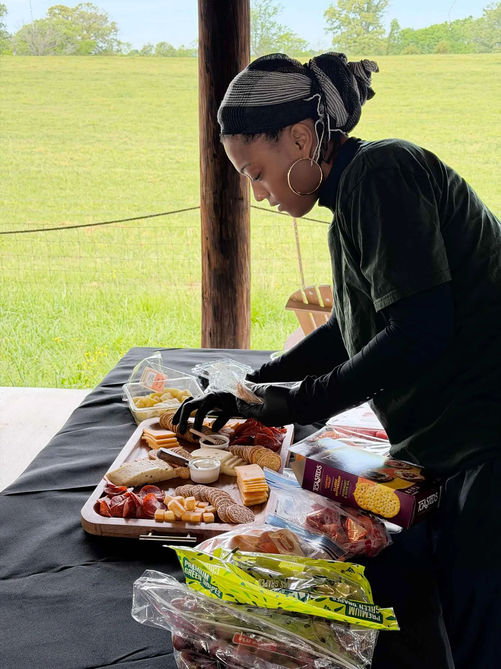 A woman with large hoop earrings and braided hair wearing a headscarf, black long sleeves, is preparing a cheese and charcuterie platter at an outdoor setting with a grassy field and blue sky in the background.