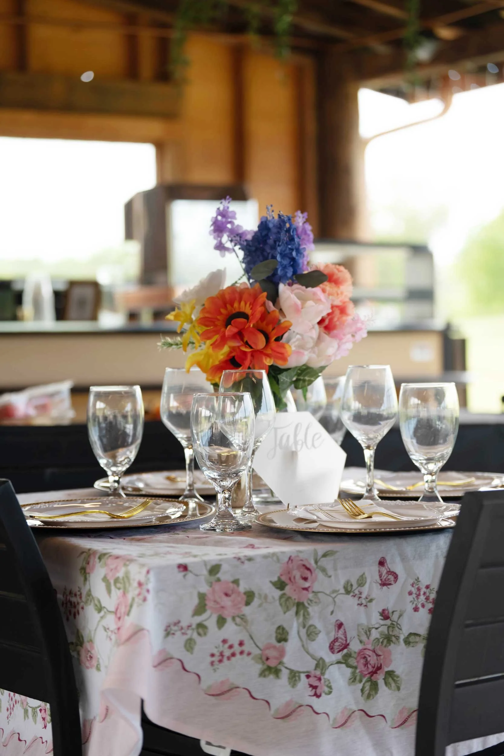 A table set for a celebration with a floral centerpiece featuring pink, purple, yellow, and blue flowers. The table has floral tablecloth and is bordered with glassware, plates, gold utensils, and a white card labeled 'Table.' The background shows a 