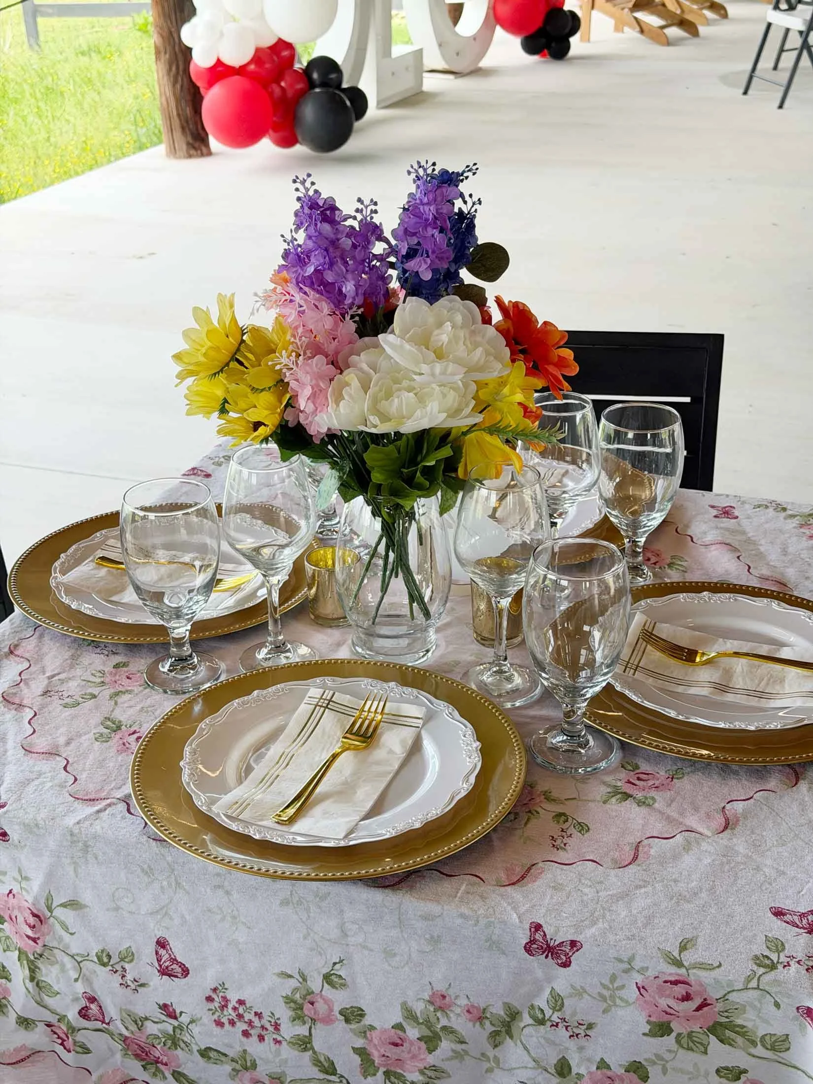 A dining table set with a floral centerpiece, gold-accented plates, gold utensils, and wine glasses on a floral tablecloth. In the background, there are balloon decorations in red, black, and white.