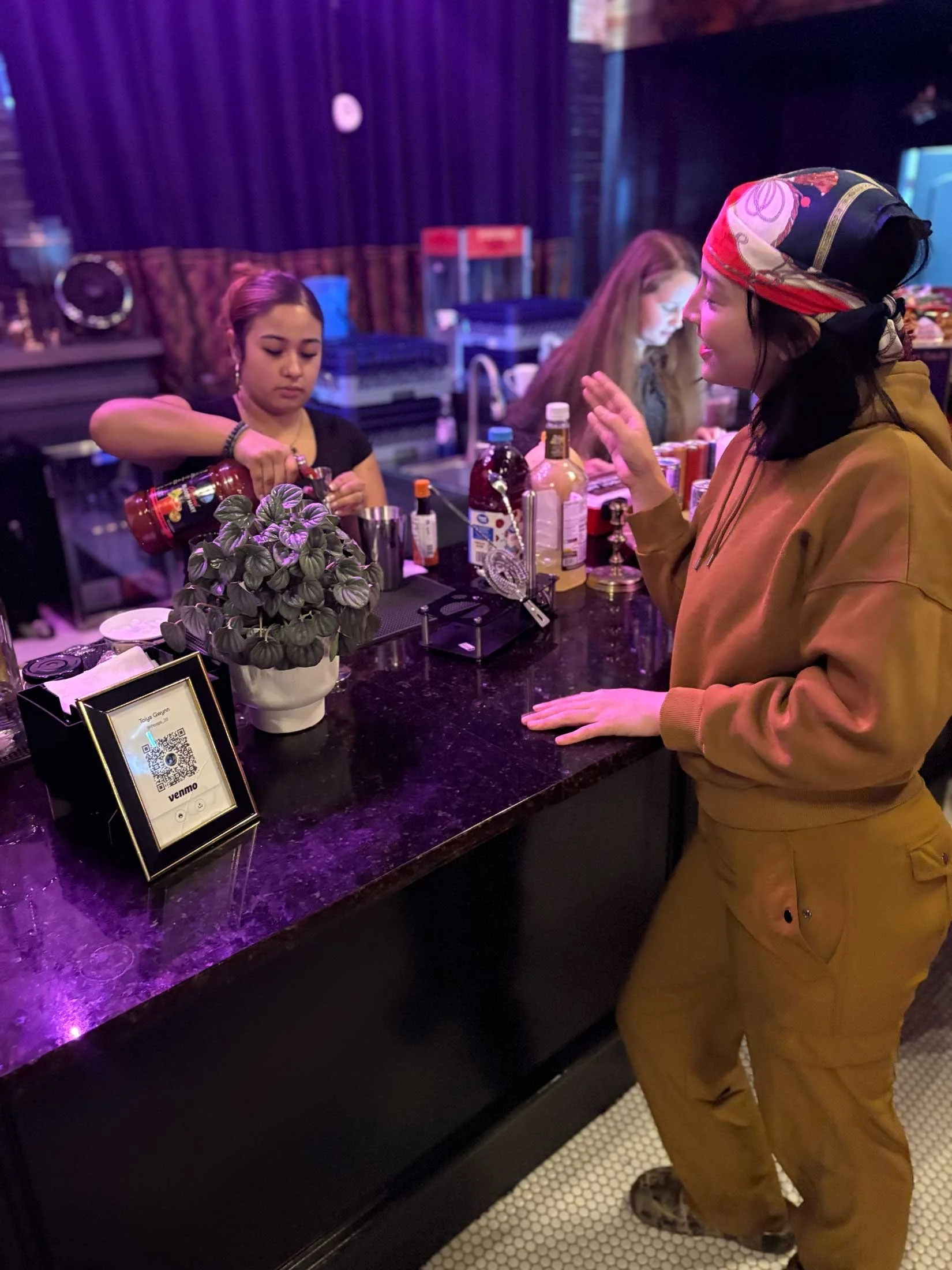 A woman with a colorful headscarf talking to a bartender behind a bar counter with bottles, a plant, and a QR code display, inside a dimly lit bar or nightclub.
