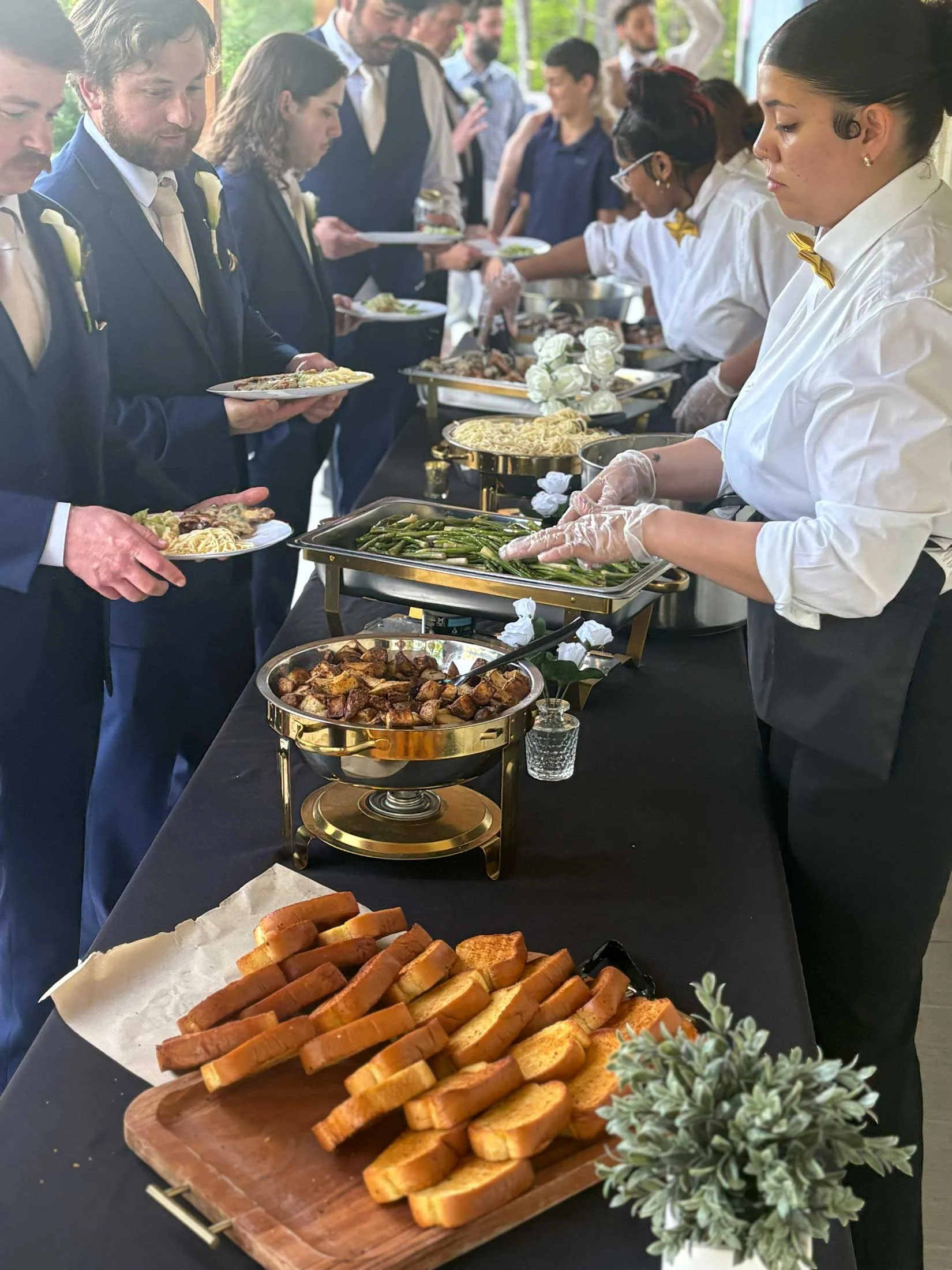 People in formal attire at a buffet line with food trays, bread, and a decorative plant in the foreground.