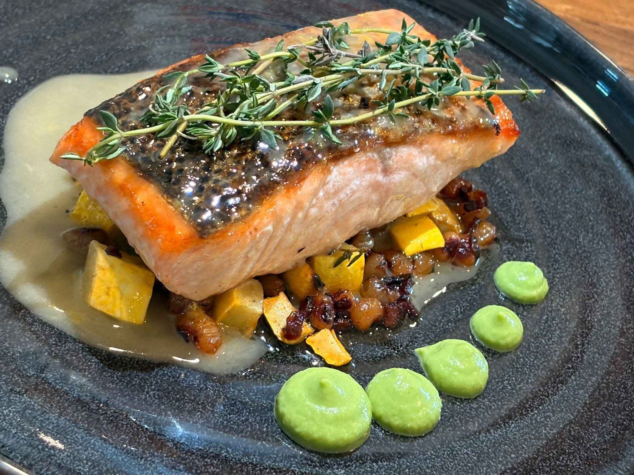 A cooked salmon filet topped with herbs, served on a black plate with roasted potatoes, vegetables, and green sauce.