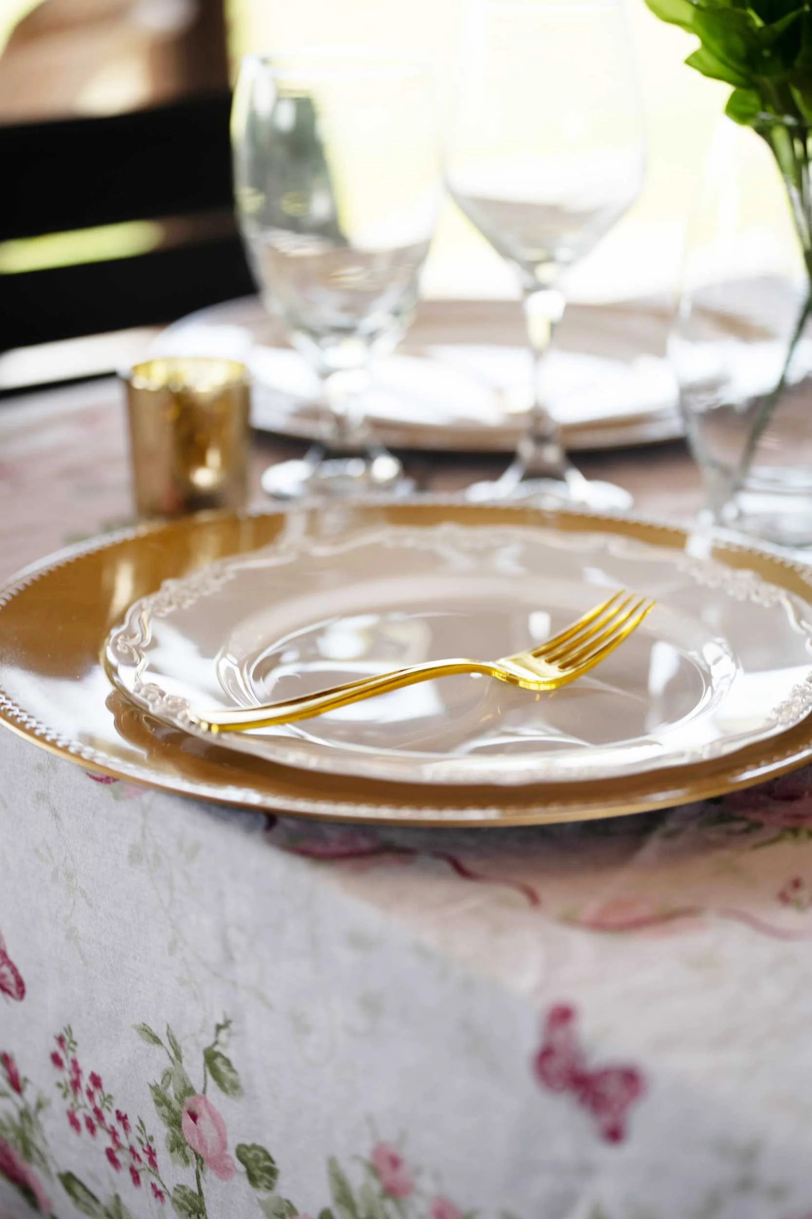 A dining table set with a gold-trimmed plate, a gold fork, and a gold cup, with wine glasses and a small vase of flowers in the background, on a floral tablecloth.
