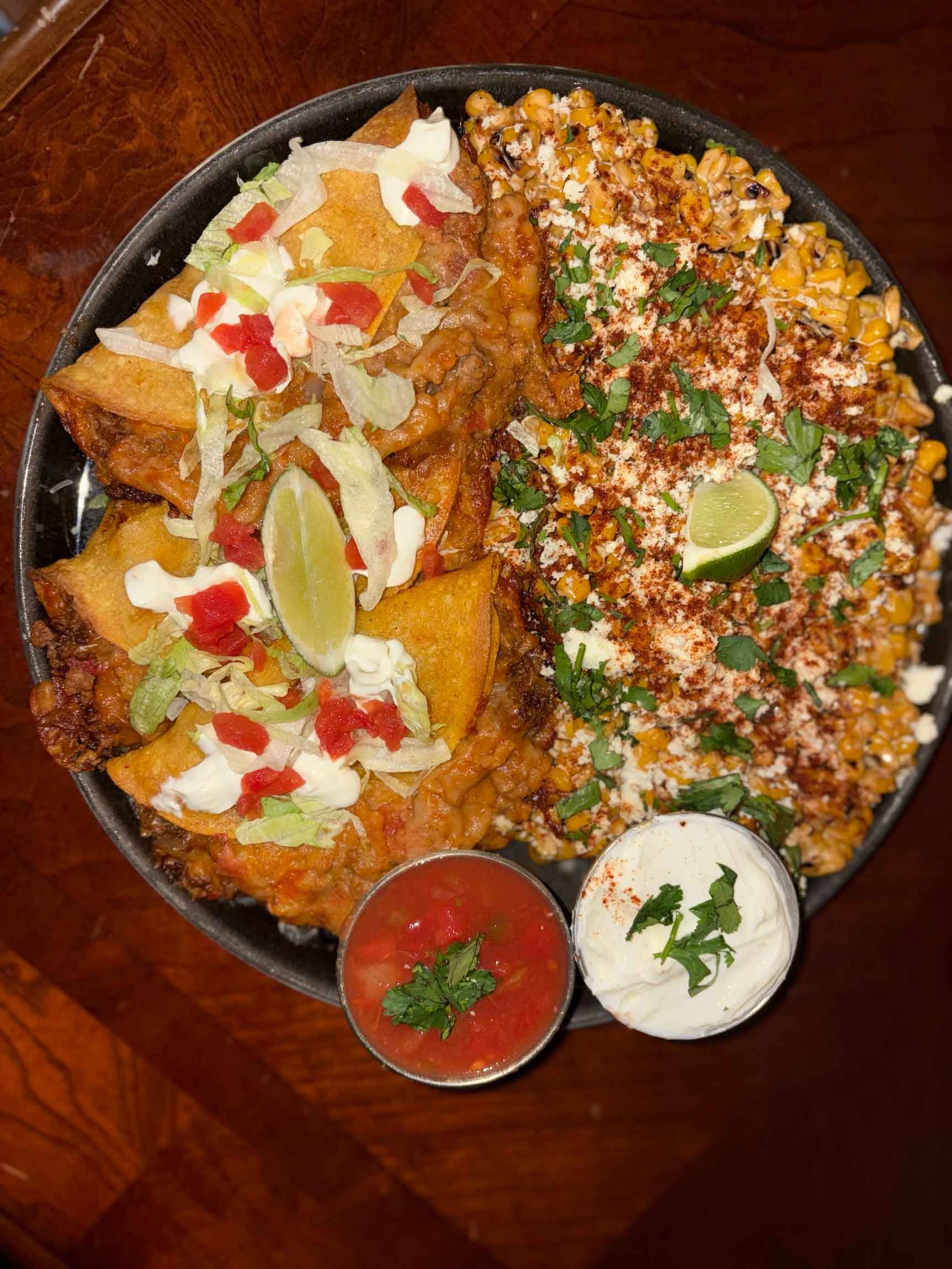 A platter of Mexican food featuring nachos with melted cheese, ground beef, lettuce, tomatoes, sour cream, and lime wedges, alongside bowls of salsa and sour cream.