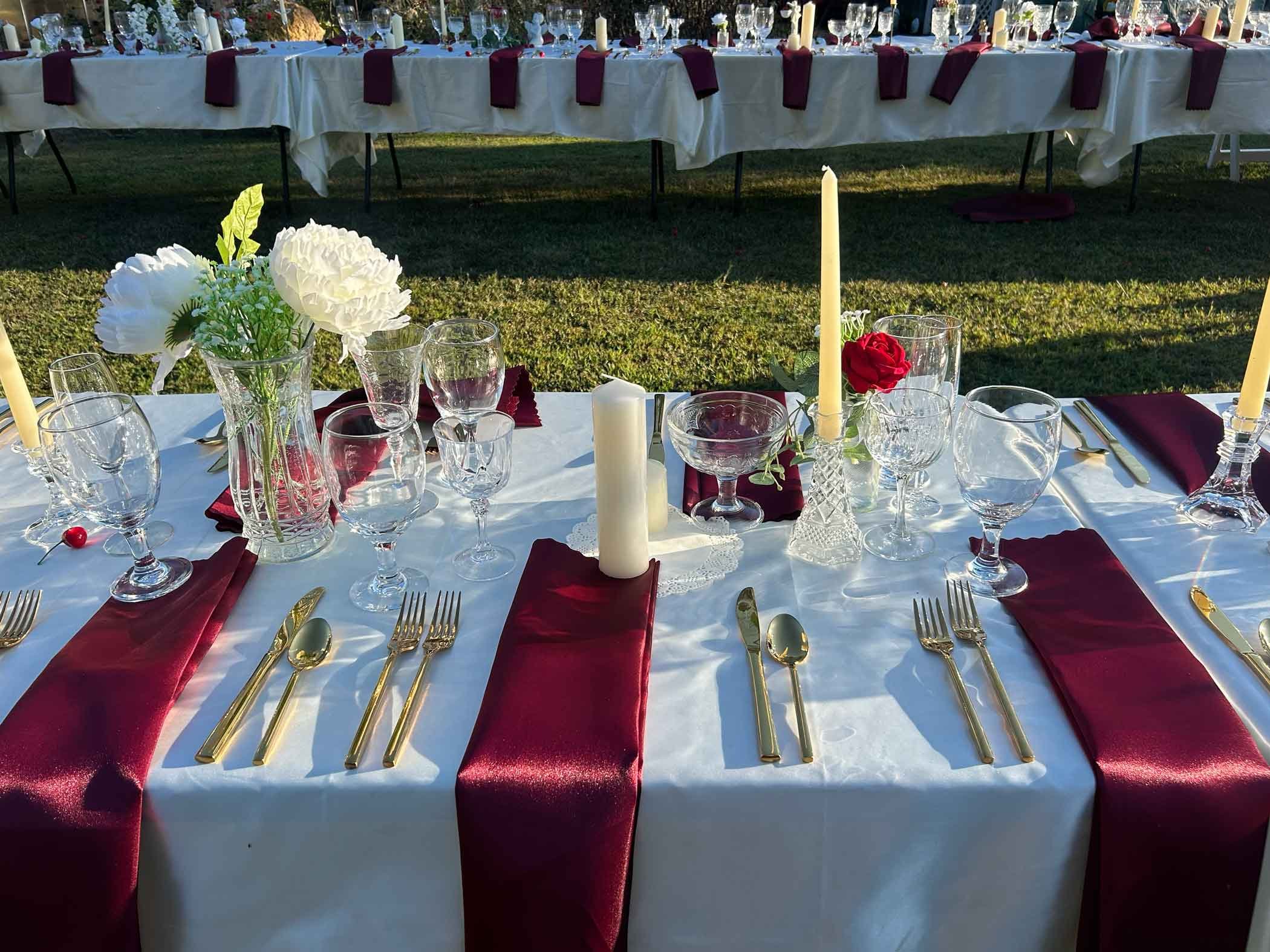 A long outdoor dining table set for an event, decorated with white tablecloths, tall candles, and floral centerpieces in glass vases. Each place setting has gold utensils, wine glasses, and maroon napkins. In the background, there is another table wi