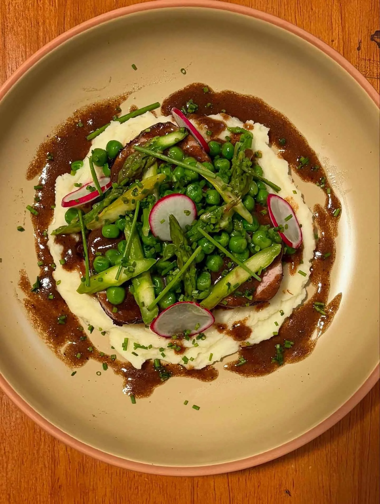 A dish of mashed potatoes topped with green peas, radish slices, and green herbs, served with gravy surrounding the mashed potatoes on a beige plate.