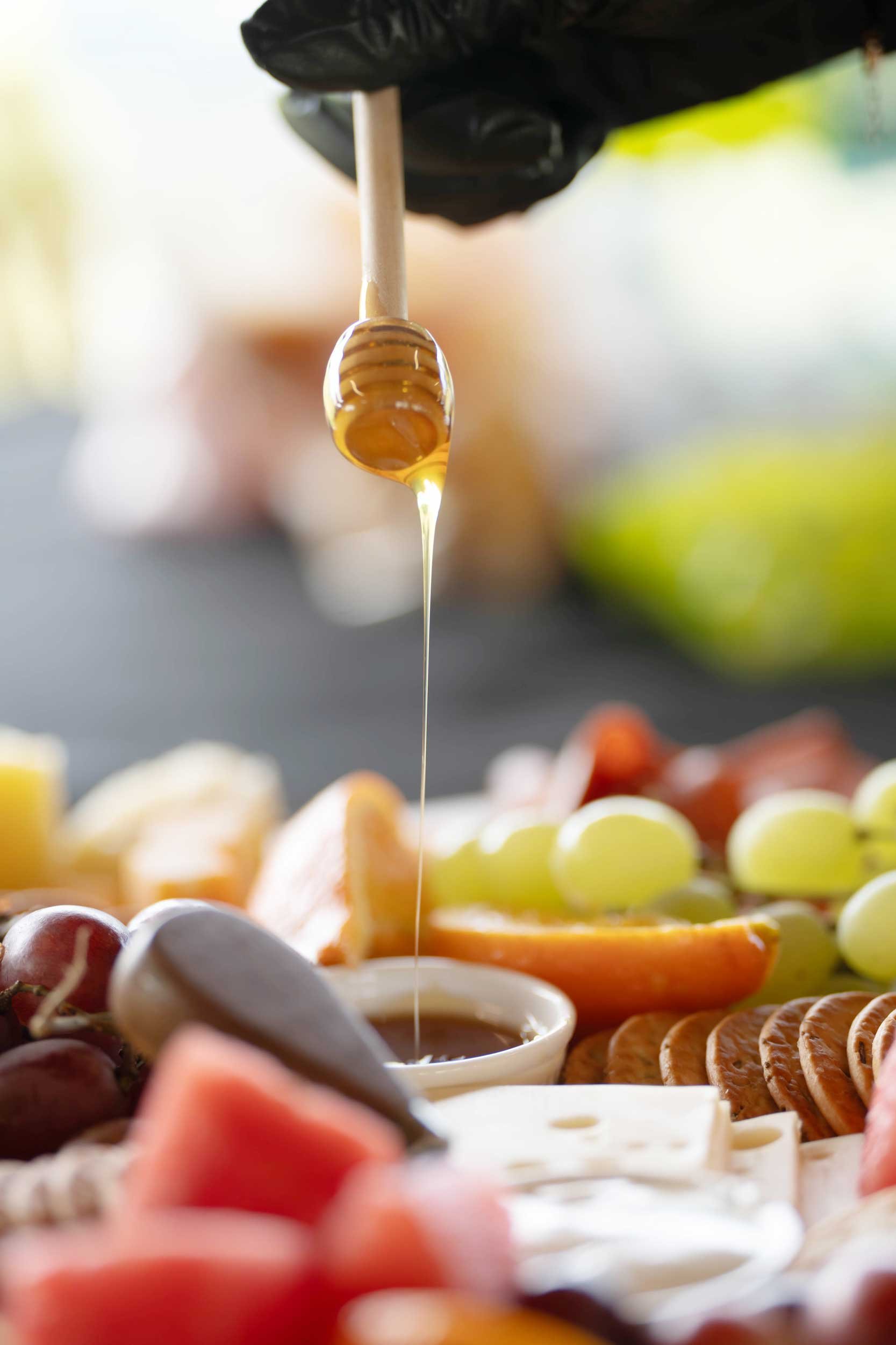 Close-up of honey being drizzled from a honey dipper over a variety of fruits and cheeses on a platter.