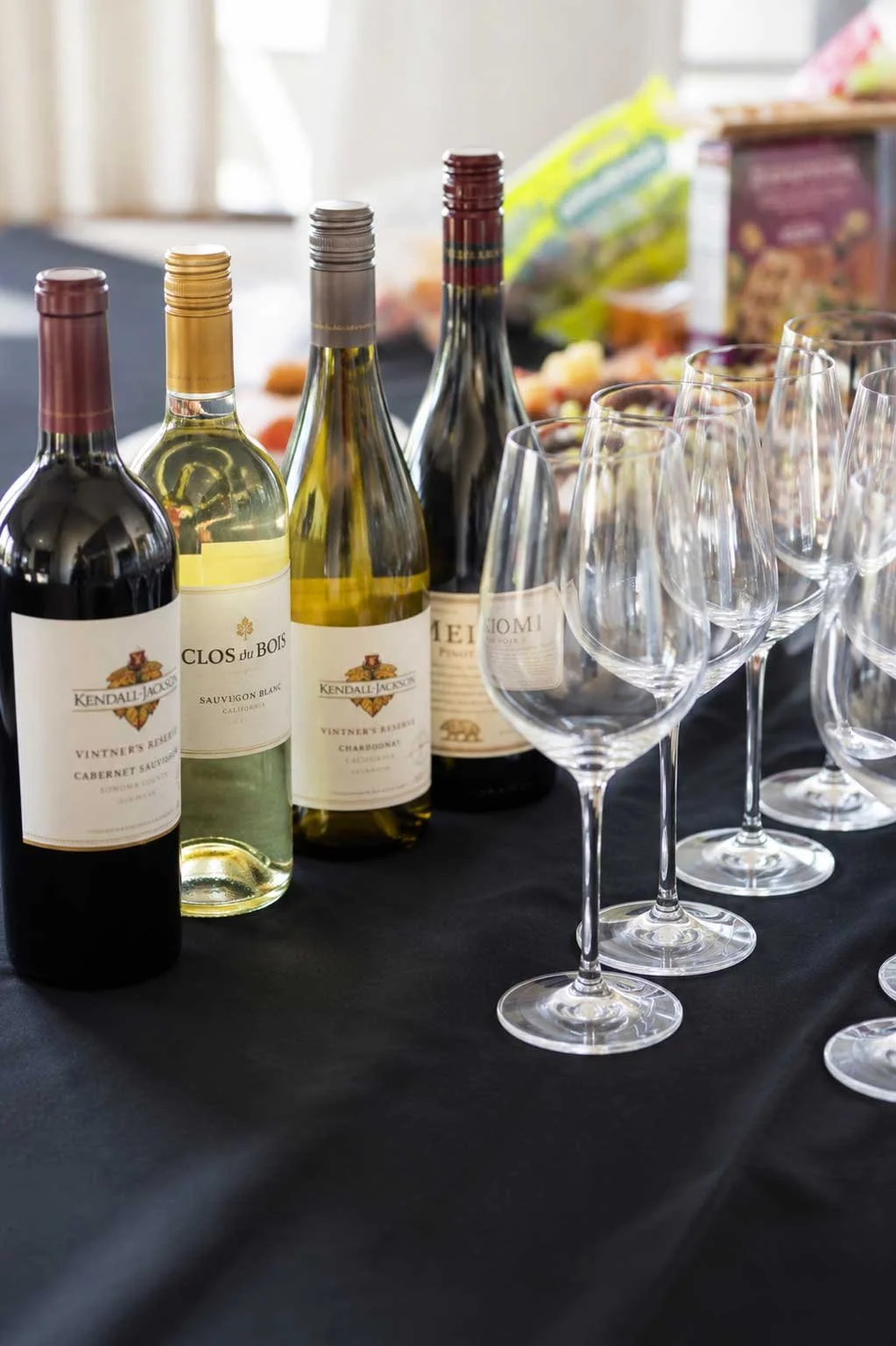 Four bottles of wine with empty wine glasses on a black tablecloth, blurred food items, and snacks in the background.