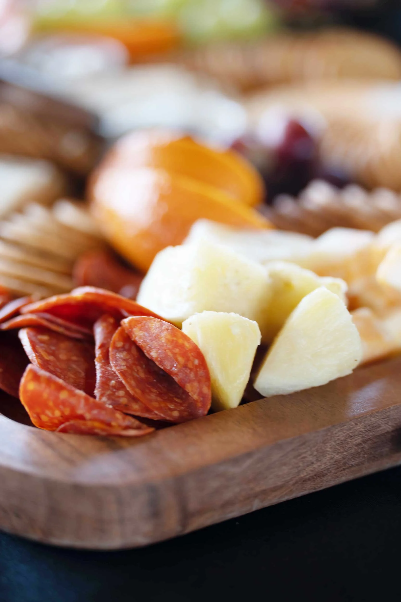 Close-up of a wooden serving board with slices of pepperoni, chunks of white cheese, and some grapes in the background.