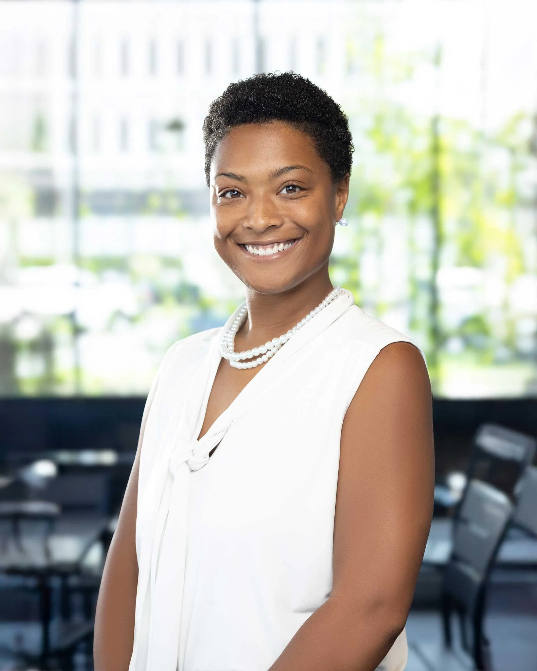 A woman smiling, wearing a white top, pearl necklace, pearl earrings, and short curly hair, with a blurred modern office background.