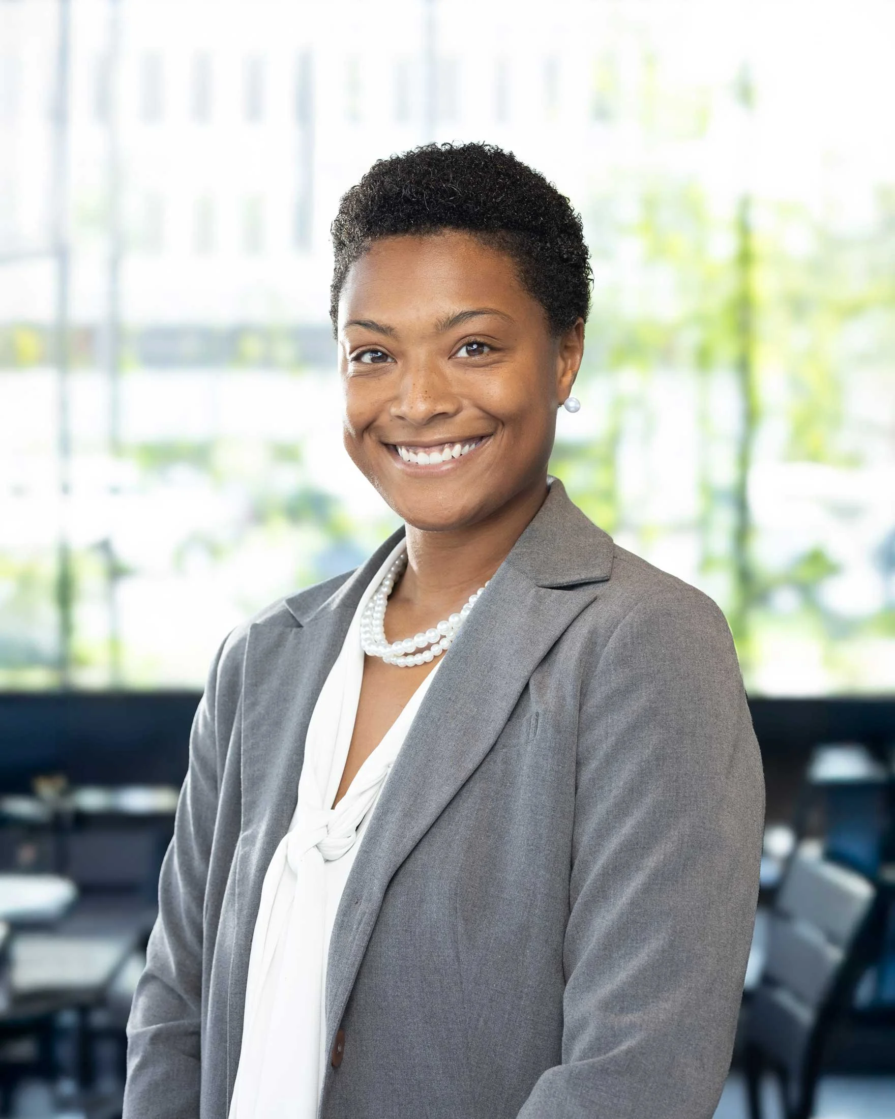 Headshot of a smiling African American woman in business attire, wearing a gray blazer, white blouse, pearl necklace, and pearl earrings, standing in a bright, modern office environment.