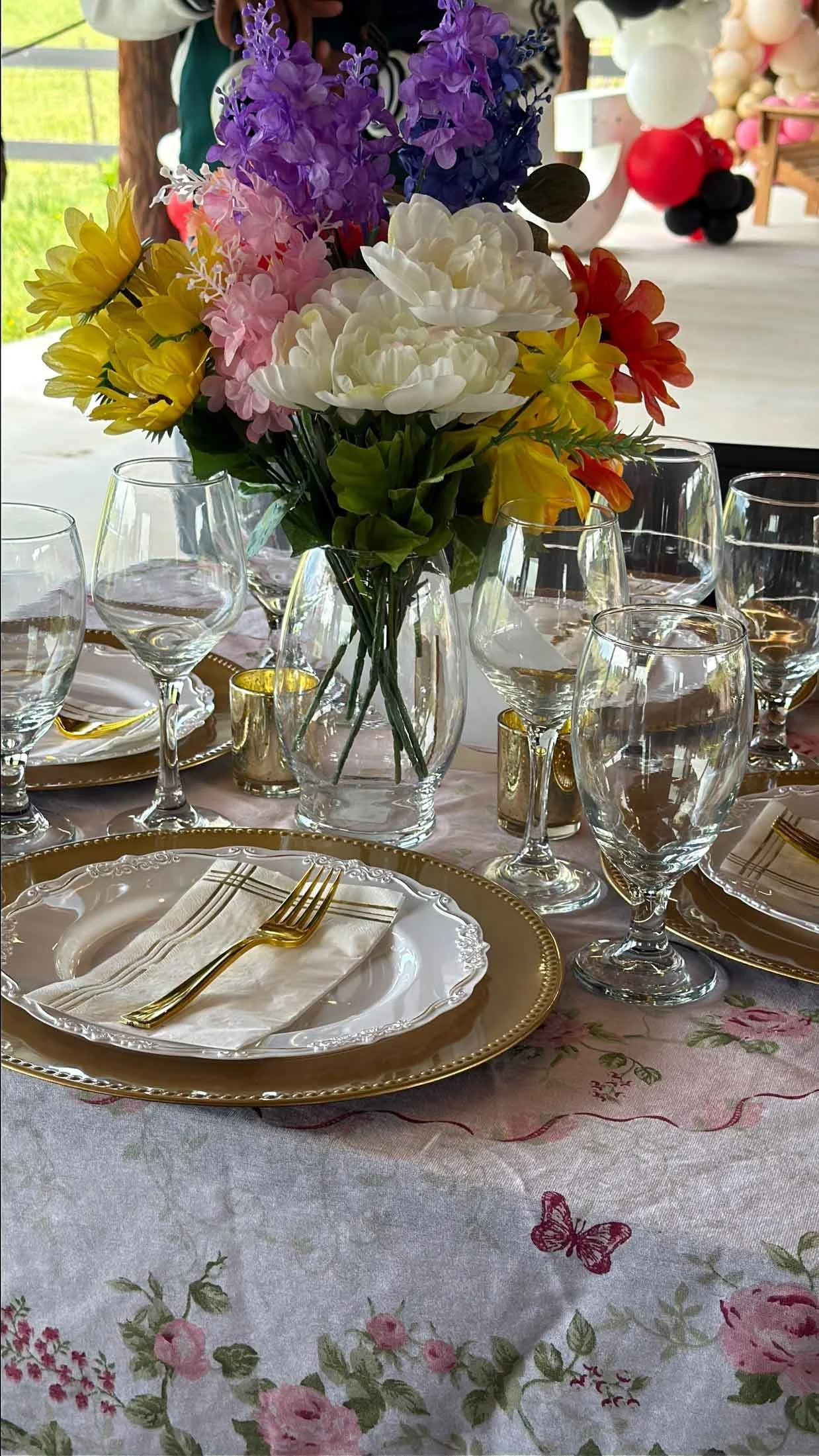 Table setting with a floral centerpiece of white, purple, yellow, pink, and orange flowers, surrounded by clear wine glasses, and gold-trimmed plates with napkins and gold utensils, on a floral tablecloth.
