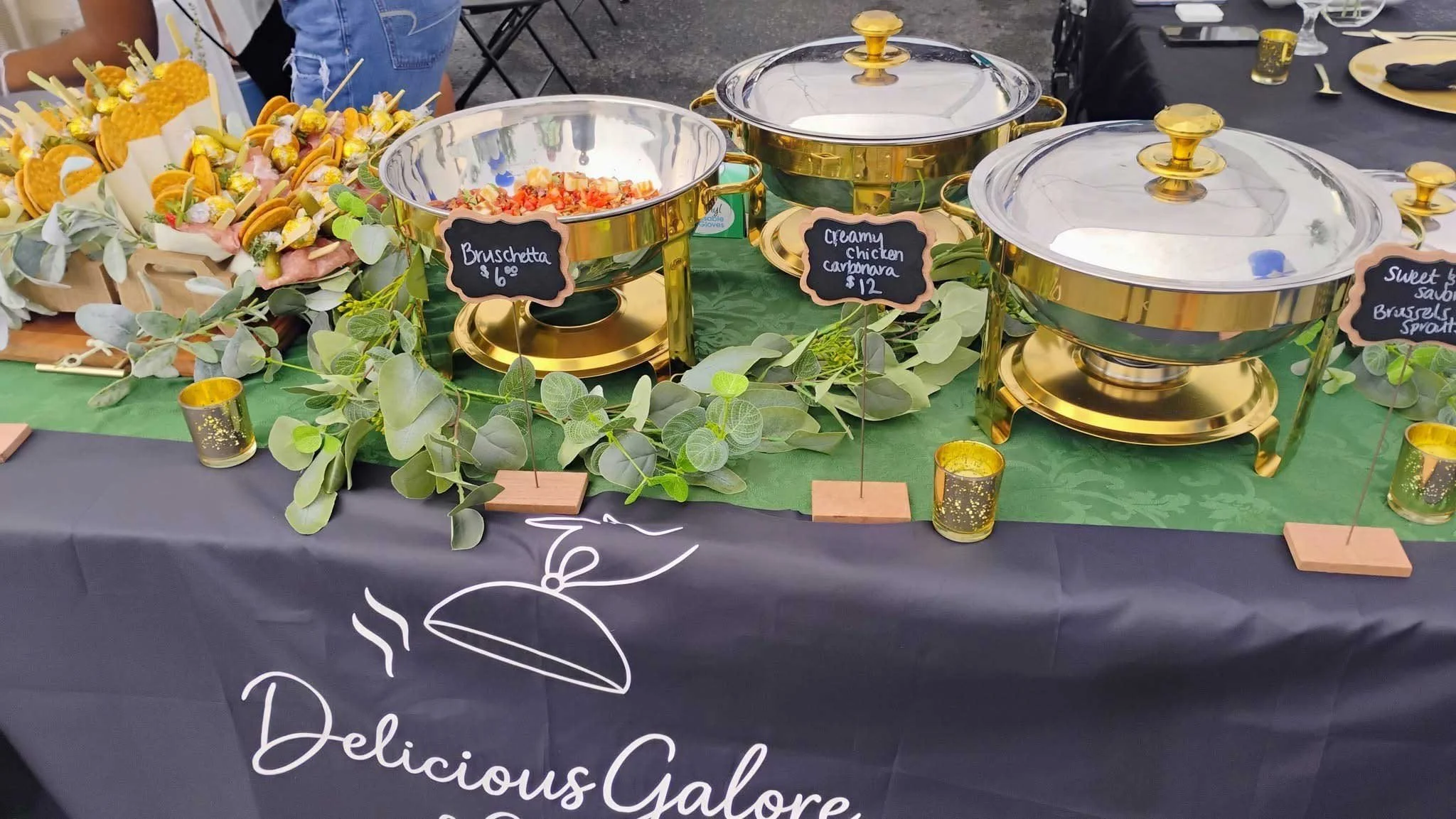 A buffet table at a food event with three gold chafing dishes covered with glass lids, a separate large stainless steel bowl with salad, and a tray of sandwiches with pickles and skewers, all decorated with green leaves. Small chalkboard signs label 