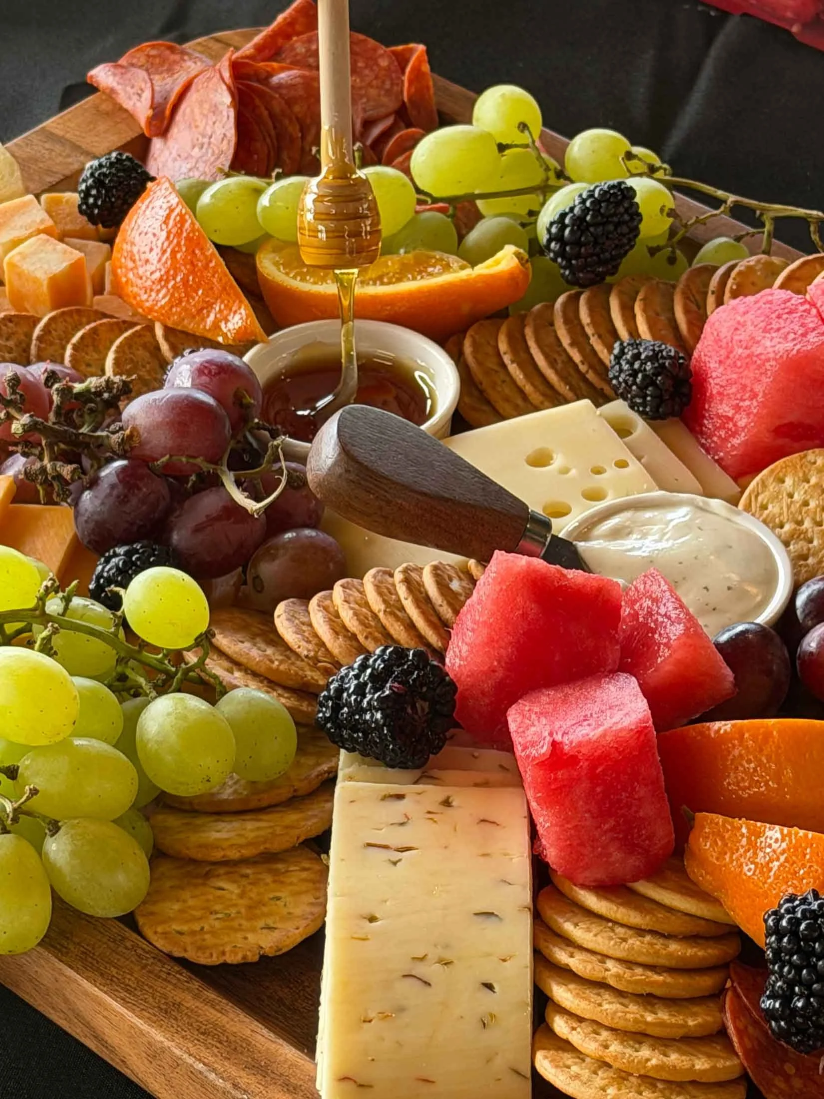 A wooden charcuterie board with various cheeses, grapes, sliced fruits like watermelon, orange, and strawberry, crackers, sliced cured meats, blackberries, and honey drizzled from a honey dipper.